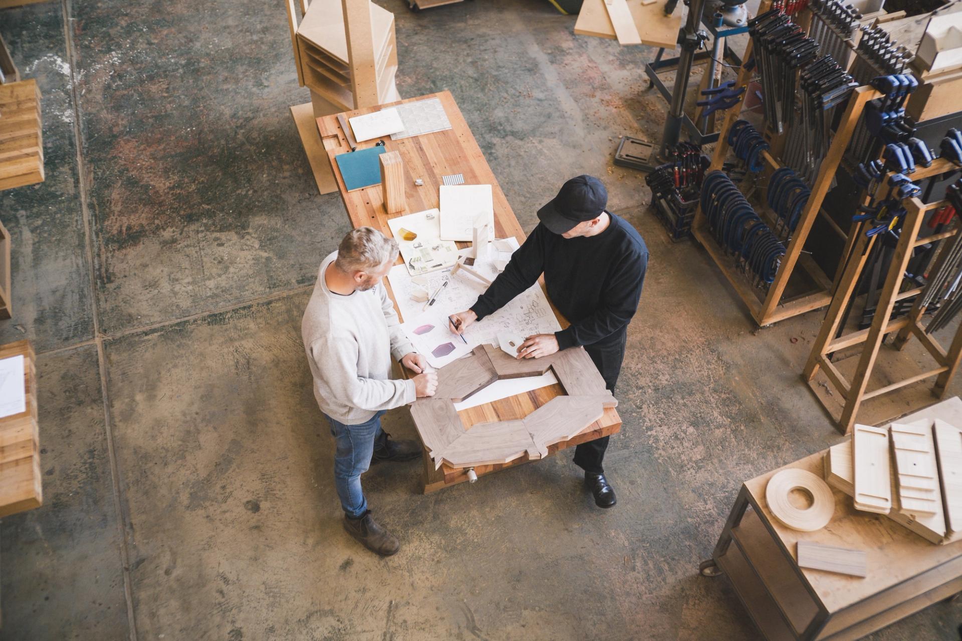 2 men standing on either side of a workbench engaged in conversation. The workbench is covered with hand-drawn design sketches.
