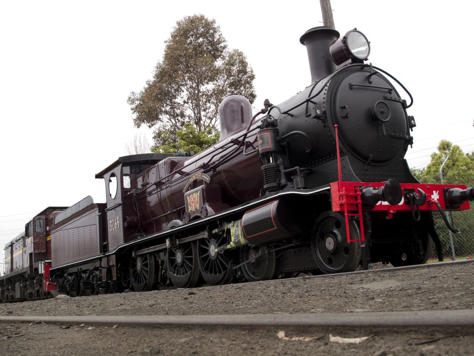 Stationary shiny black locomotive with dark green carriages outside a large brick building with schoolgirls in uniform looking on.