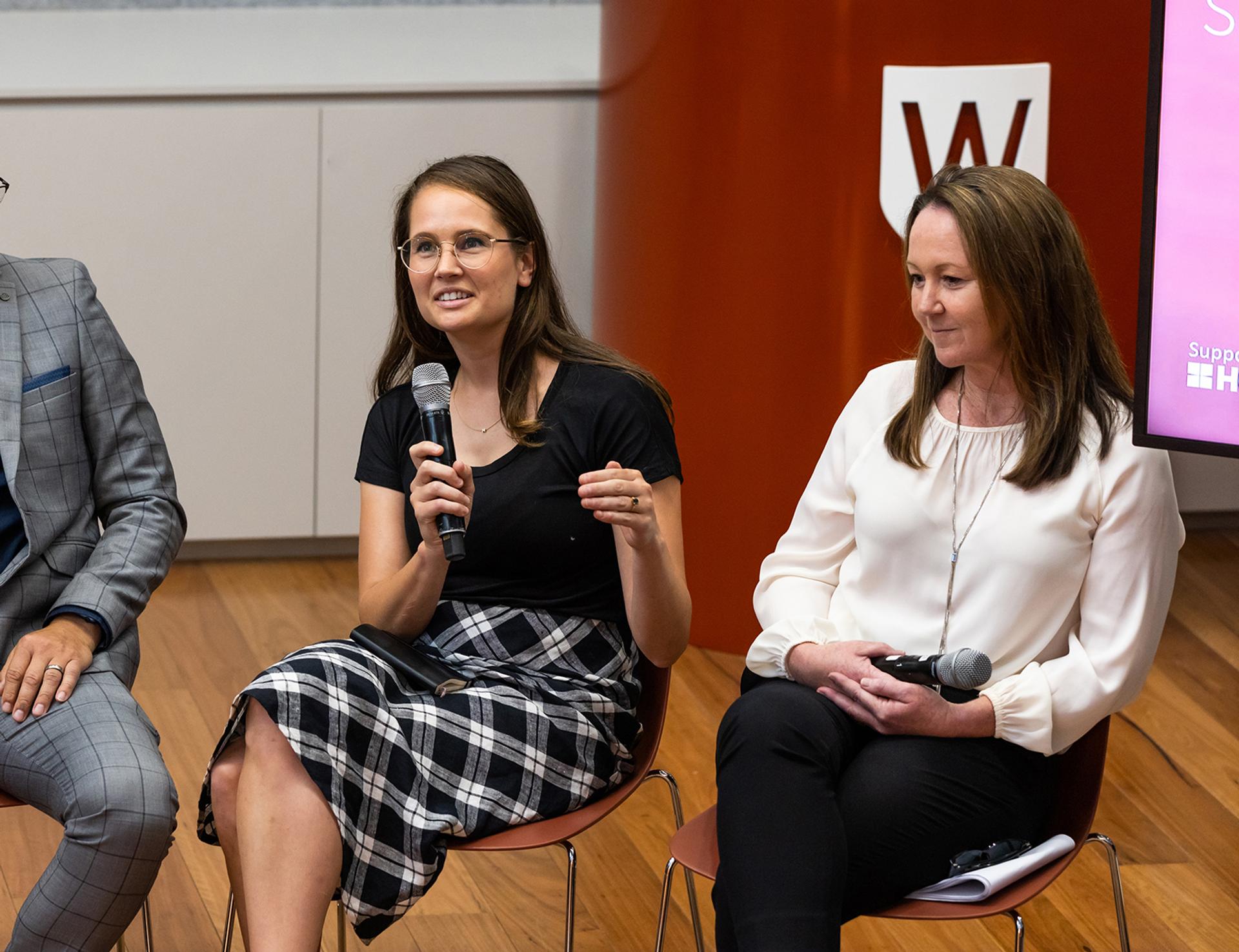 Two women sitting with microphones in their hand, at a panel.