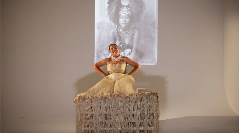 A young woman wearing traditional Samoan dress sitting on a bark covered box. There is an image of a Samoan high chief’s daughter projected behind her.