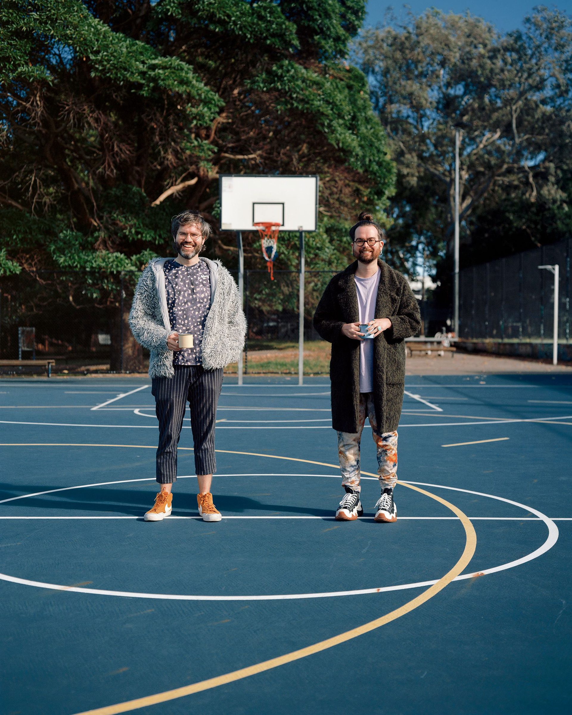 Two people stand on a blue basketball court. They are smiling at the camera and holding mugs in their hands.