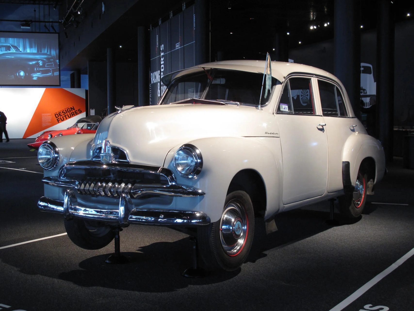 Photo of a light grey, beige car in a dark showroom. A 1955 FJ225 Holden Special four door sedan featuring a pale grey exterior with chrome brightwork. The interior of the car comprises red leather upholstery for the seats, two-toned red and pale grey for the inner door trim, a red painted dashboard and red carpeted floor at the back.