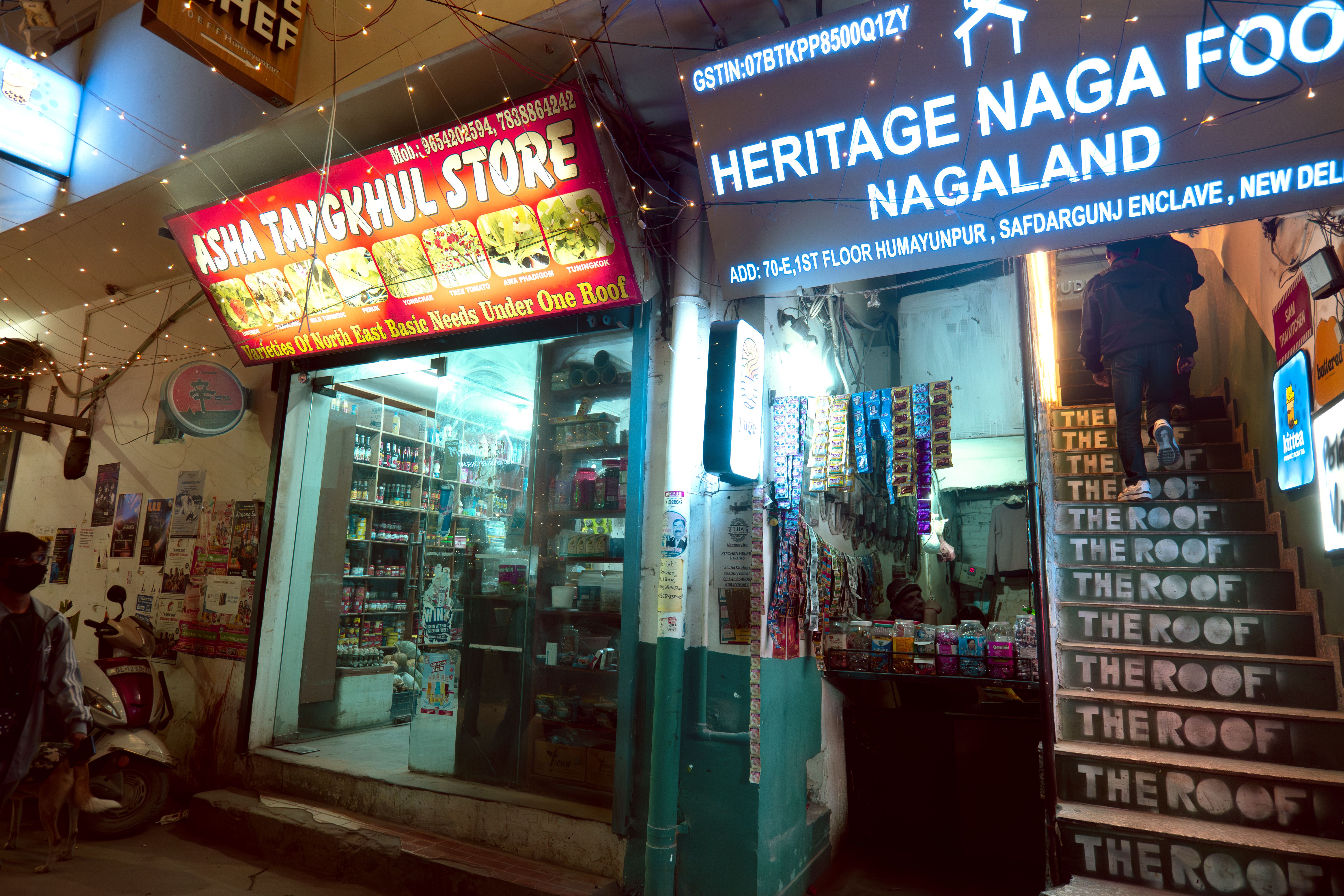 Two small grocery stores with lightbox signage. On the left, ‘ASHA TANGKHUL STORE’ in white on a red background with 8 images of produce. On the right ‘HERITAGE NAGA FOOD NAGALAND’ in white on a black background.