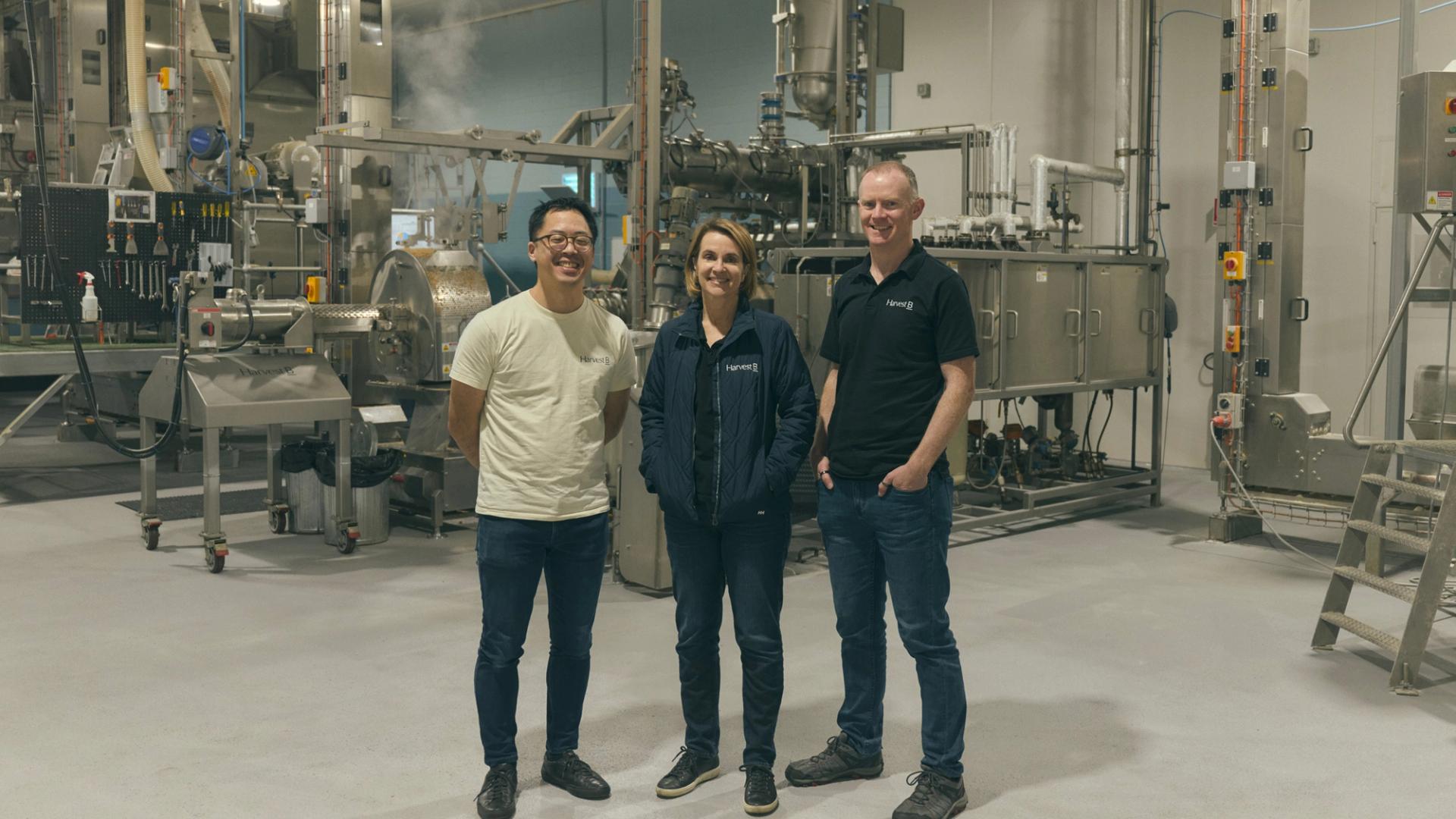 Three people stand together in an industrial food processing facility, surrounded by large metal machinery and equipment. They are dressed casually, with two of them wearing branded shirts and one in a light jacket. All three are smiling as they pose for the camera, conveying a sense of teamwork and professionalism in the high-tech, clean environment.