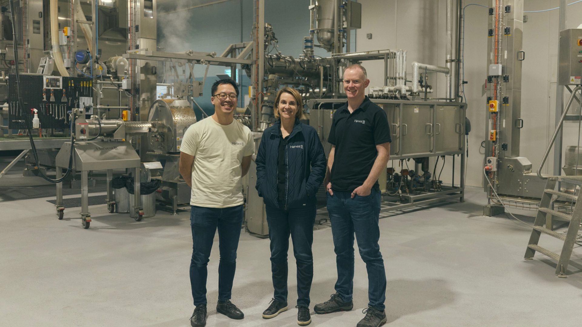 Three people stand together in an industrial food processing facility, surrounded by large metal machinery and equipment. They are dressed casually, with two of them wearing branded shirts and one in a light jacket. All three are smiling as they pose for the camera, conveying a sense of teamwork and professionalism in the high-tech, clean environment.