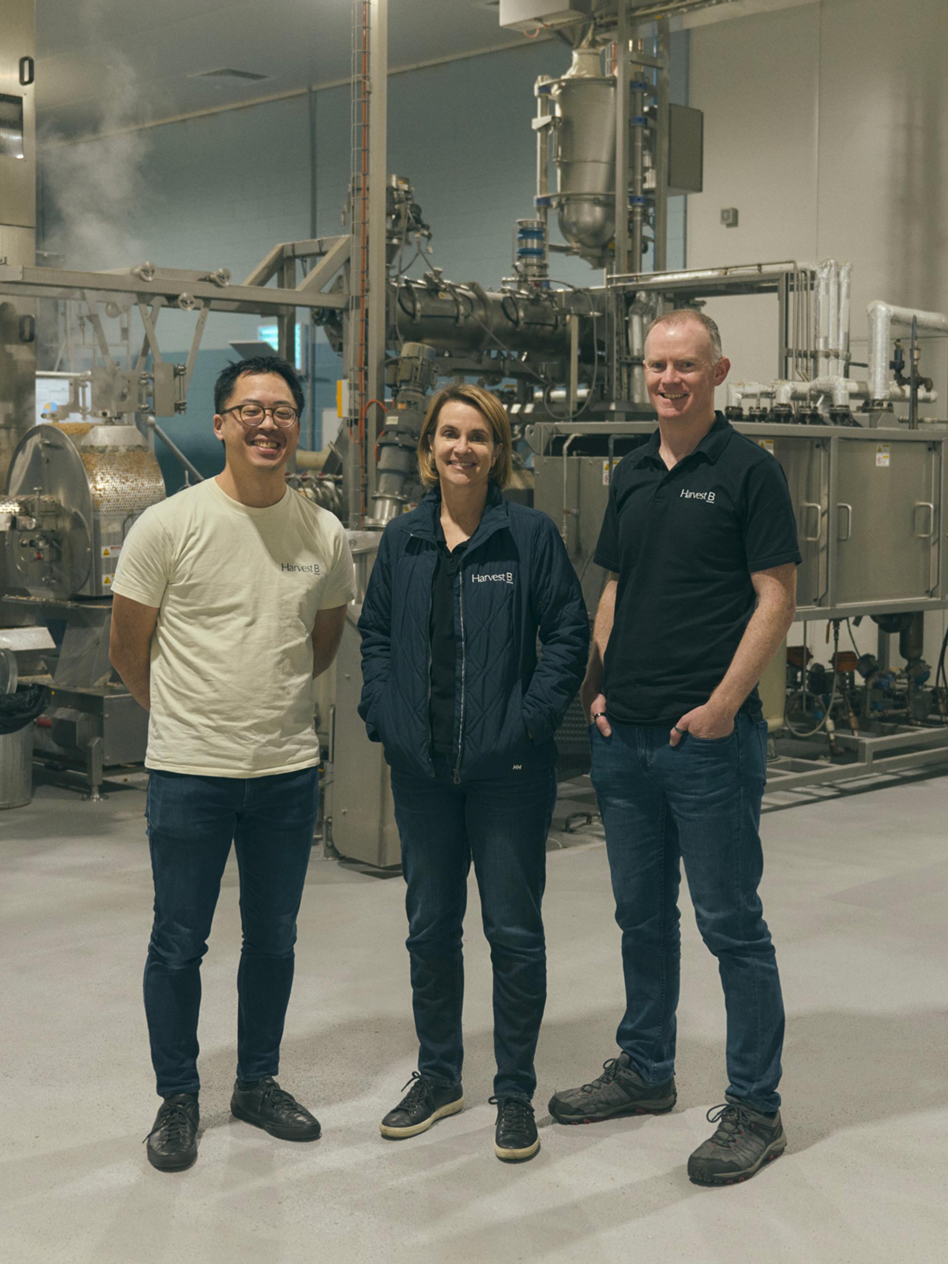Three people stand together in an industrial food processing facility, surrounded by large metal machinery and equipment. They are dressed casually, with two of them wearing branded shirts and one in a light jacket. All three are smiling as they pose for the camera, conveying a sense of teamwork and professionalism in the high-tech, clean environment.