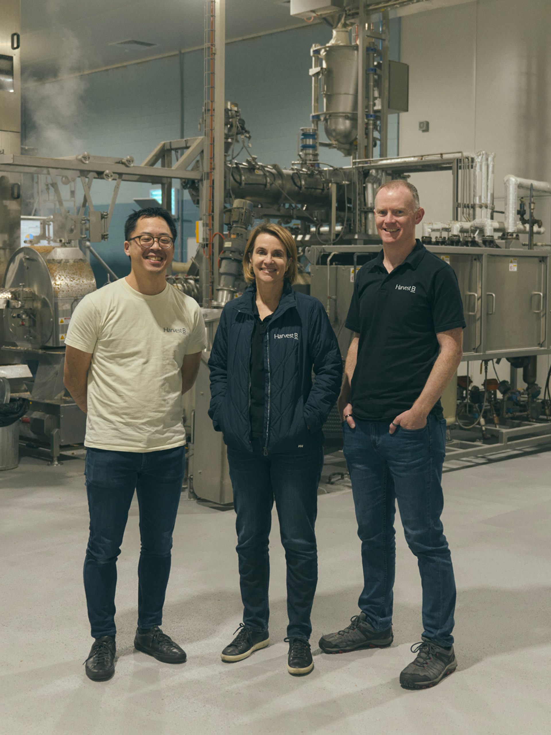 Three people stand together in an industrial food processing facility, surrounded by large metal machinery and equipment. They are dressed casually, with two of them wearing branded shirts and one in a light jacket. All three are smiling as they pose for the camera, conveying a sense of teamwork and professionalism in the high-tech, clean environment.