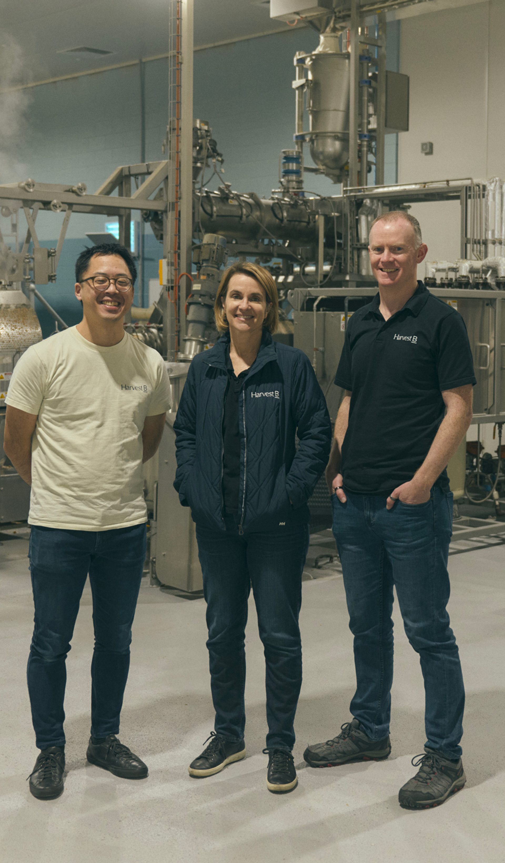 Three people stand together in an industrial food processing facility, surrounded by large metal machinery and equipment. They are dressed casually, with two of them wearing branded shirts and one in a light jacket. All three are smiling as they pose for the camera, conveying a sense of teamwork and professionalism in the high-tech, clean environment.