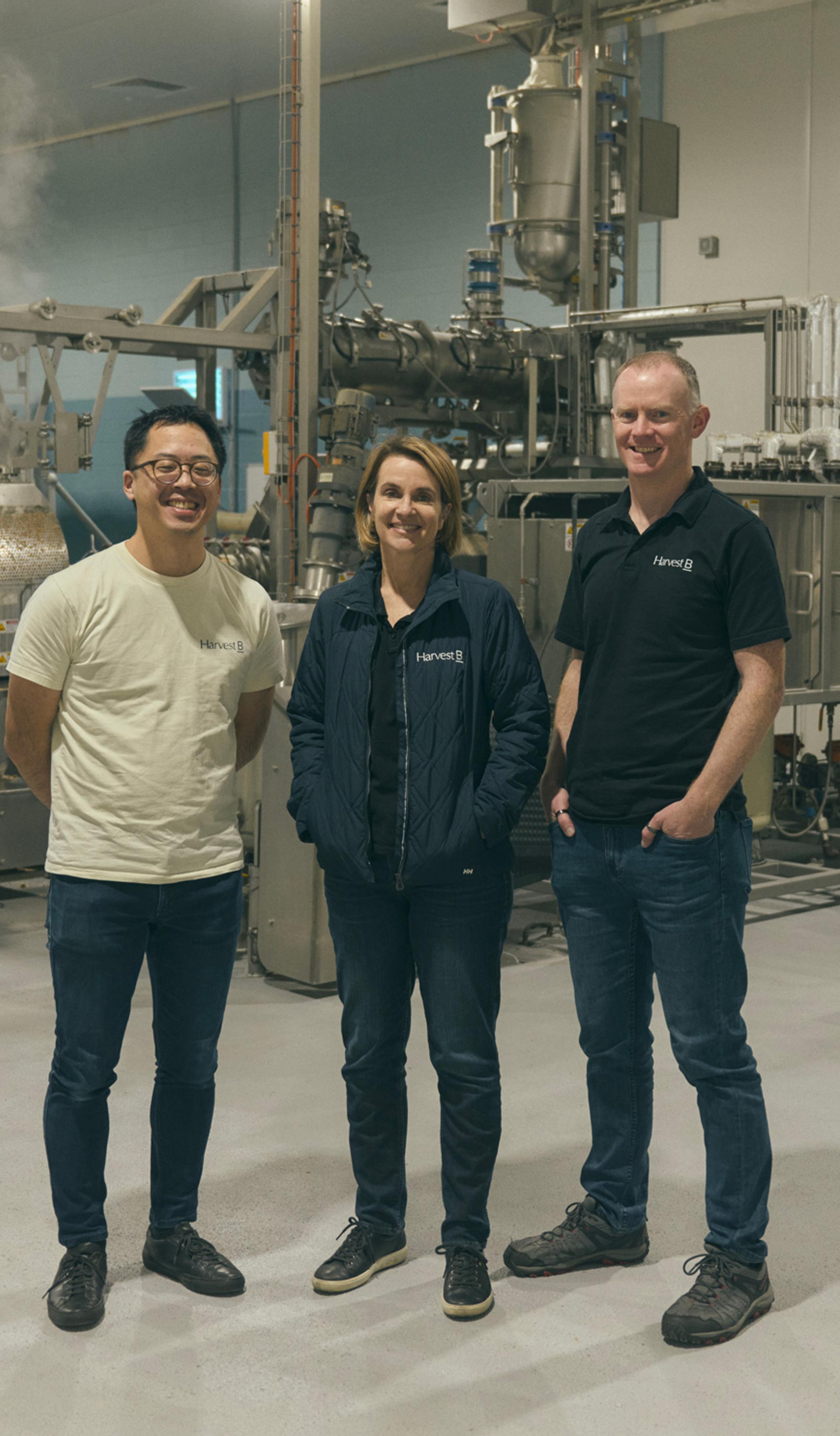 Three people stand together in an industrial food processing facility, surrounded by large metal machinery and equipment. They are dressed casually, with two of them wearing branded shirts and one in a light jacket. All three are smiling as they pose for the camera, conveying a sense of teamwork and professionalism in the high-tech, clean environment.
