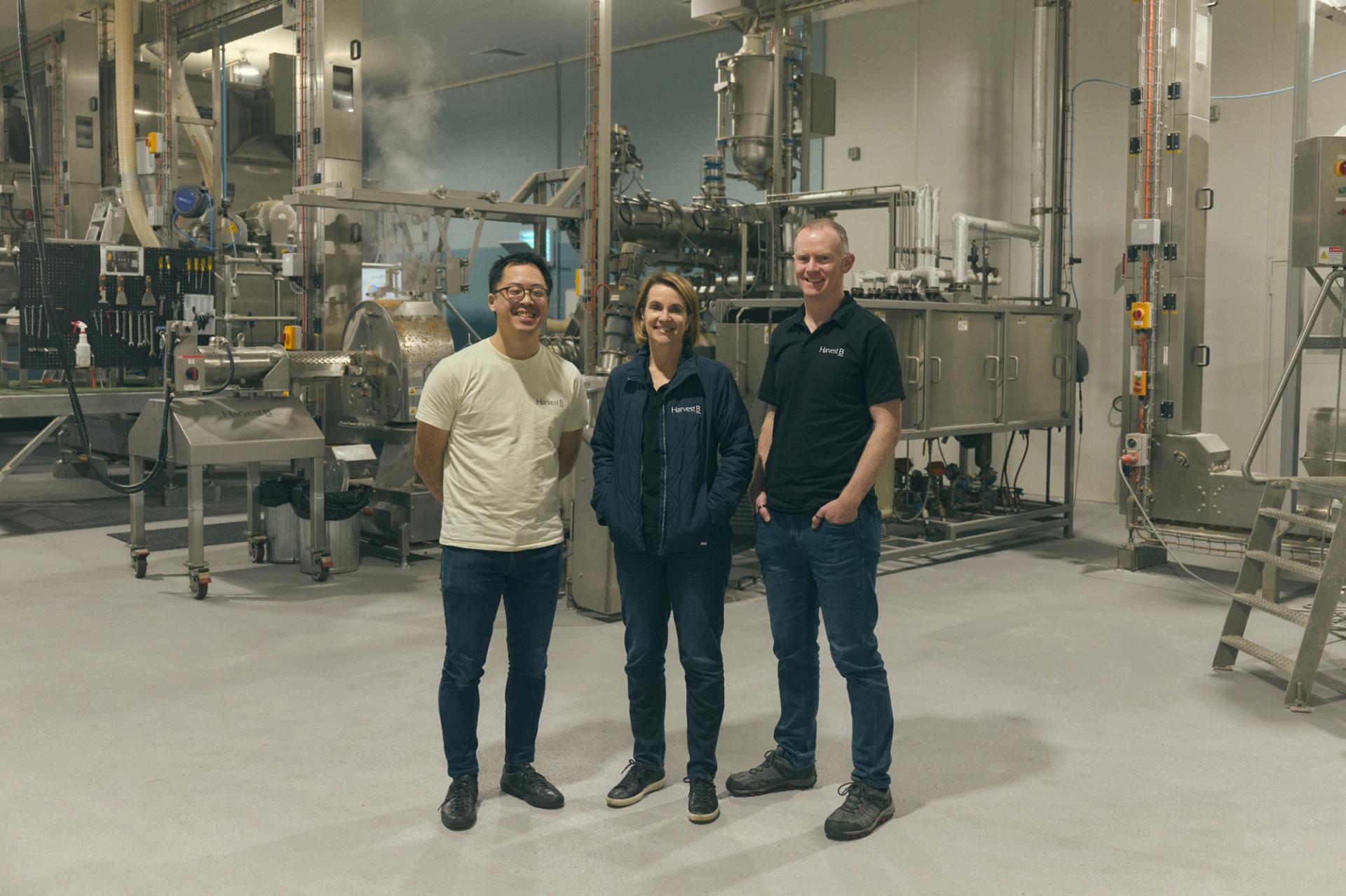Three people stand together in an industrial food processing facility, surrounded by large metal machinery and equipment. They are dressed casually, with two of them wearing branded shirts and one in a light jacket. All three are smiling as they pose for the camera, conveying a sense of teamwork and professionalism in the high-tech, clean environment.