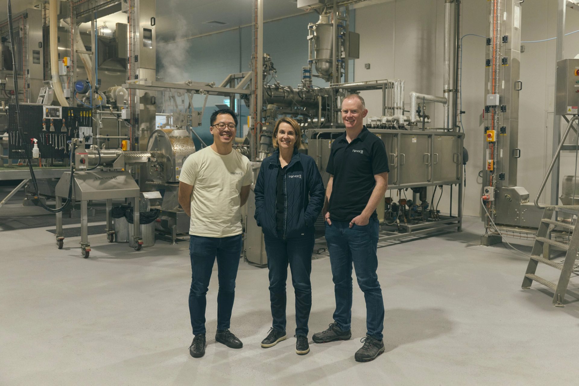 Three people stand together in an industrial food processing facility, surrounded by large metal machinery and equipment. They are dressed casually, with two of them wearing branded shirts and one in a light jacket. All three are smiling as they pose for the camera, conveying a sense of teamwork and professionalism in the high-tech, clean environment.