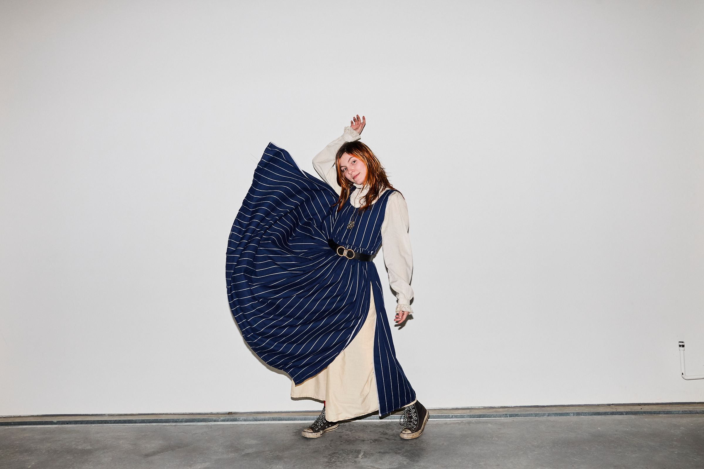 A girl posed against a white wall, throwing her blue and white striped dress to her right.