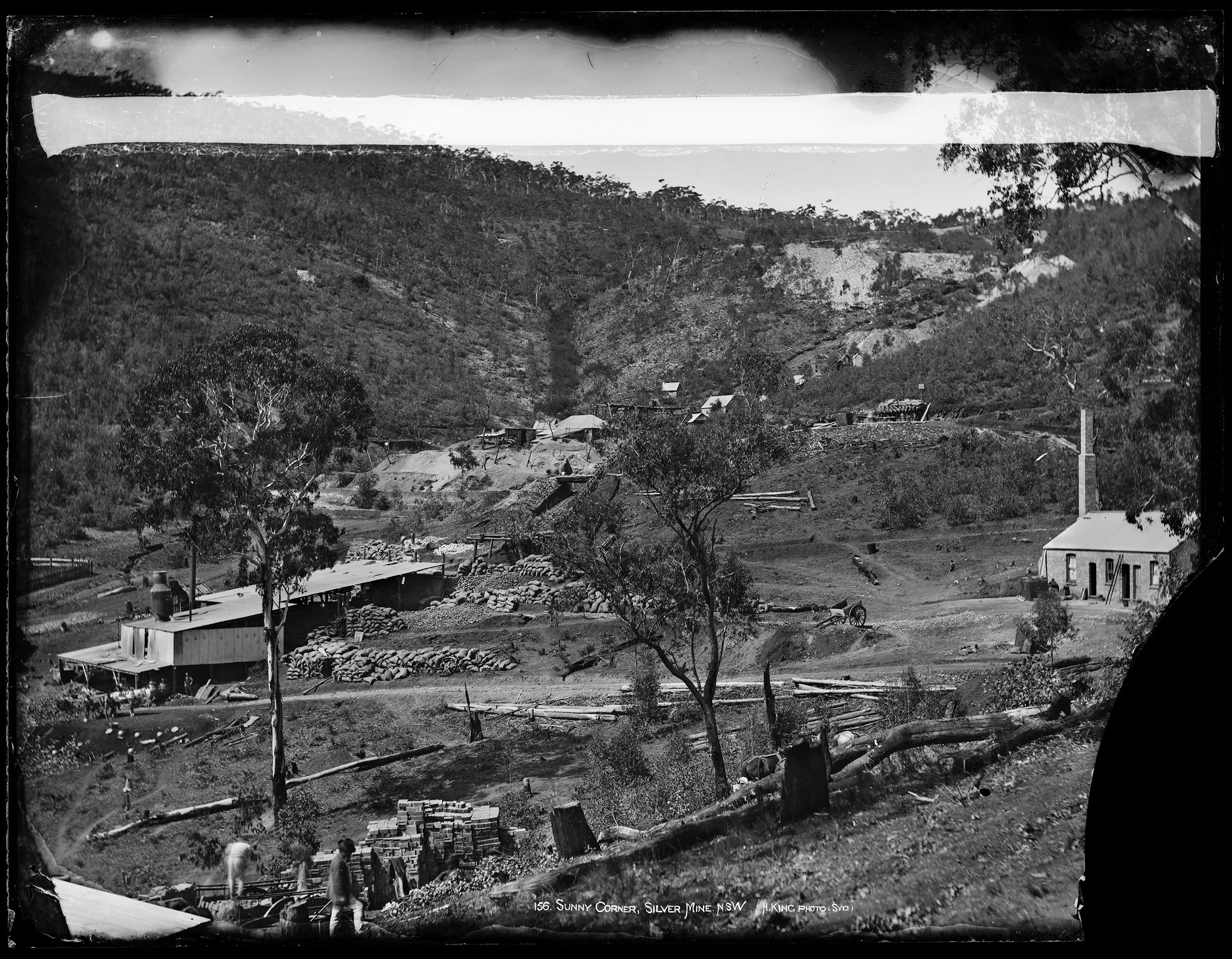 A silver mine surrounded by tall trees and some small structures. There are two men in the foreground.