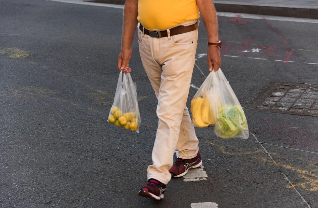 A figure wearing beige pants and a yellow shirt carries three plastic bags full of fruit.