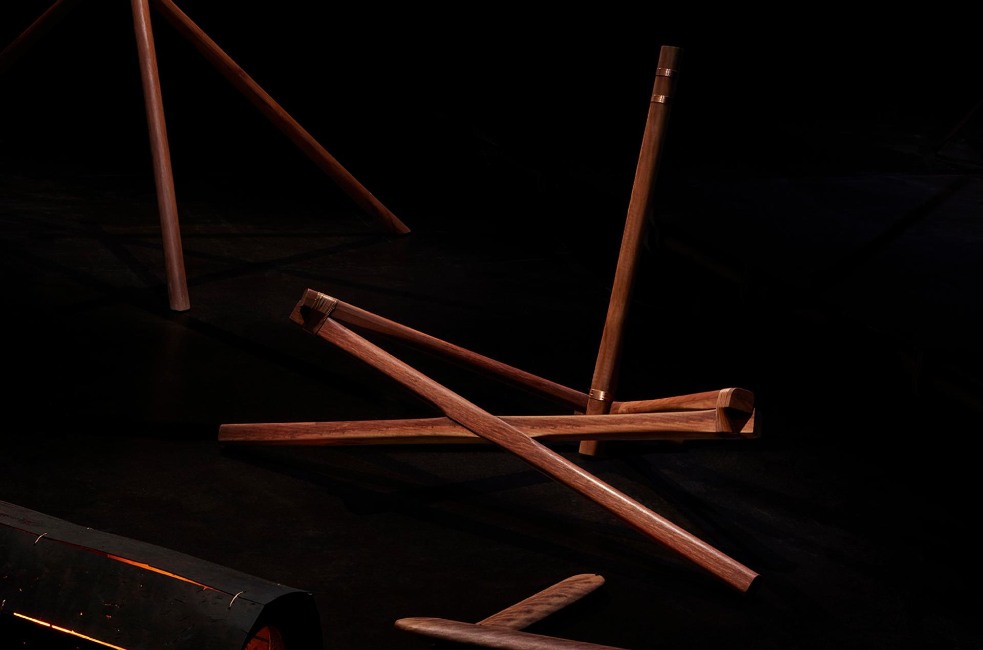 A series of interconnected dark wood beams. The sculpture is against a dark black background and is spotlit.