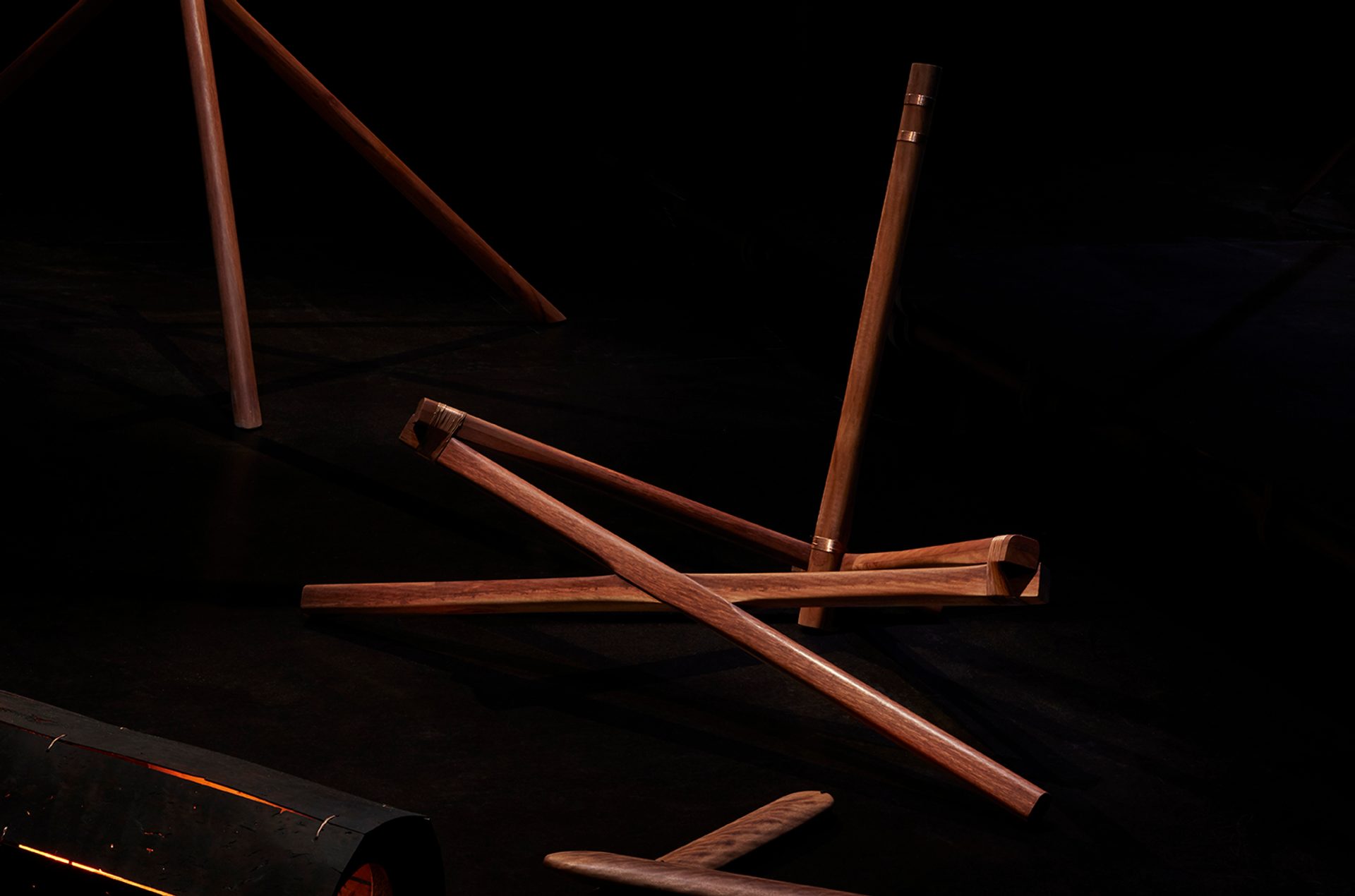 A series of interconnected dark wood beams. The sculpture is against a dark black background and is spotlit.