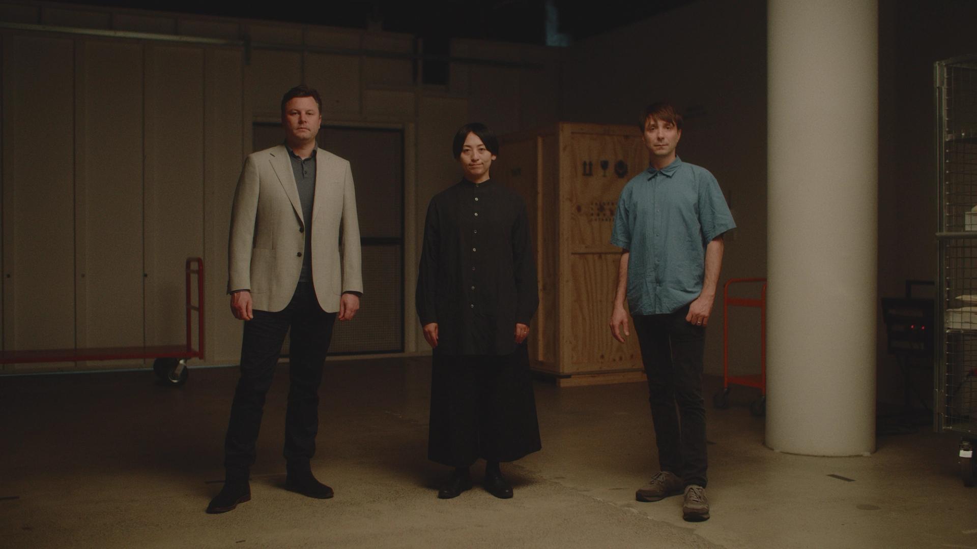 Three humans stand in a line in a garage facing the camera with muted faces