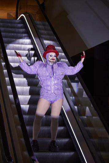 A drag king dressed in a purple sparkly suit jacket and short shorts stands on an escalator with their arms raised up. They are in the midst of a performance.