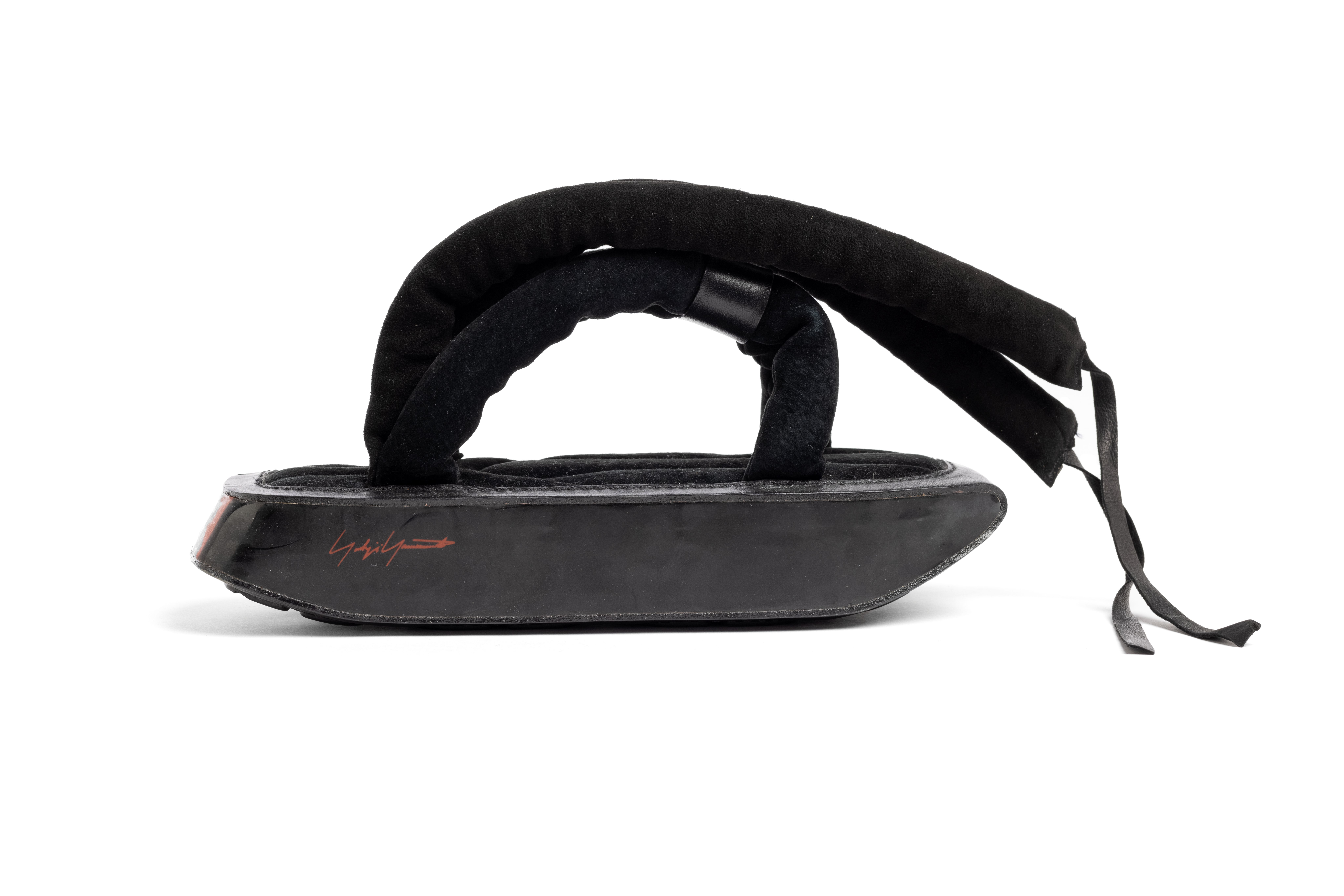 A pair of sandals with a thick flat platform synthetic rubber soles covered on the sides with aromatic polyester based thermoplastic polyurethane which is decorated with three red stripes - the Adidas mark - at the heels and Yohji Yamamoto's signature also in red on one side. The inner soles are of the black padded synthetic suede as are the straps that loop on either side of the foot and bound together with a strip of black leather. There are two more black padded straps that wind around the ankles and tie with short black leather straps.