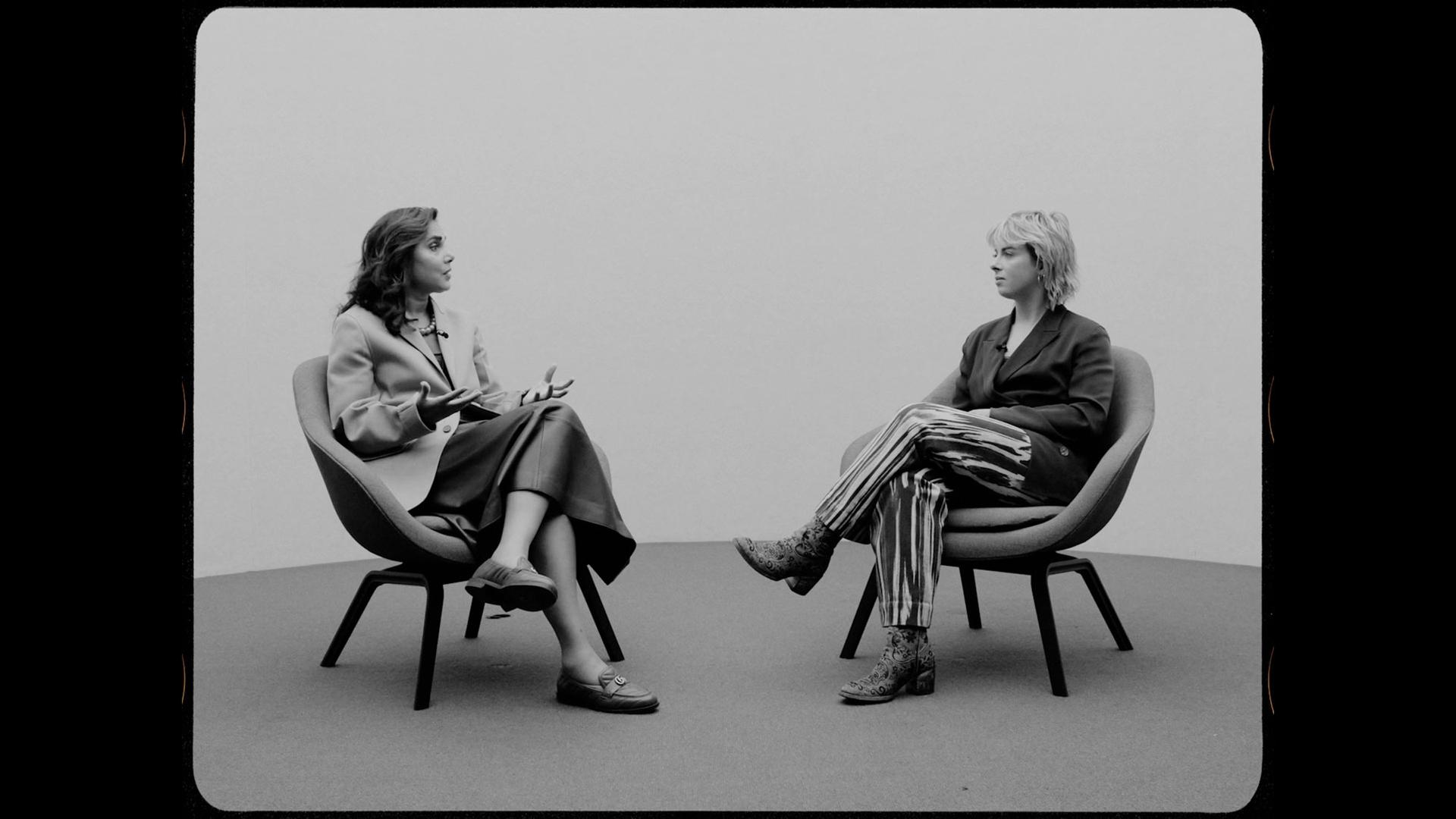 Two women sit on chairs on a stage talking to each other.