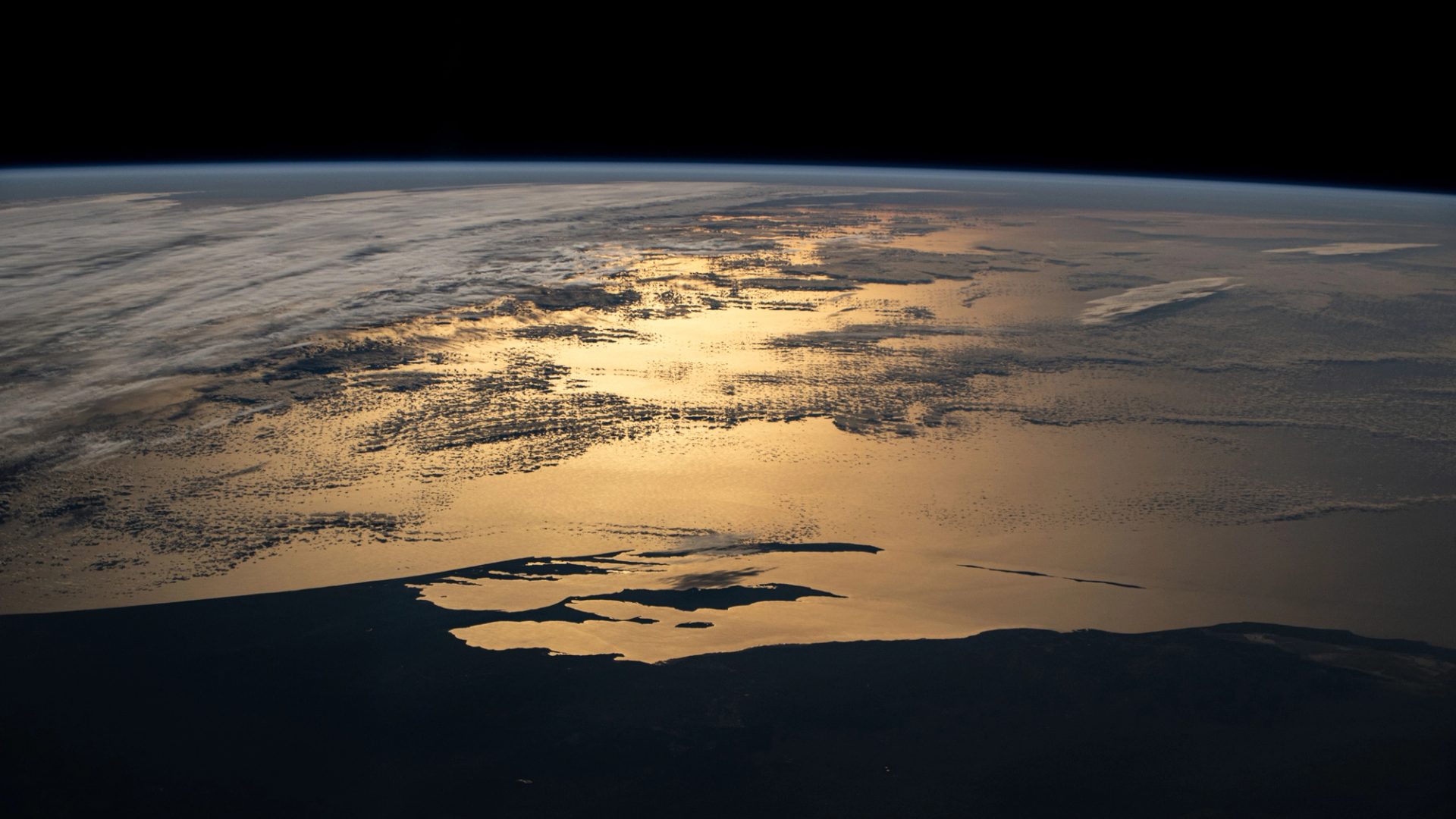 Photography of an orbital sunset taken from the International Space Station showing Western Australia's silhouetted coastline, contrasting the sun's golden-hued glint beaming off a partly cloudy Indian Ocean.