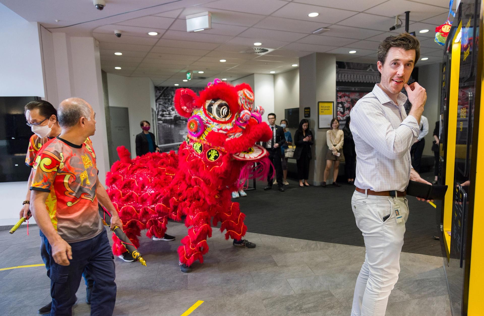A Lunar New Year lion is in the foyer of a building, a figure in a business shirt smiles in the foreground.