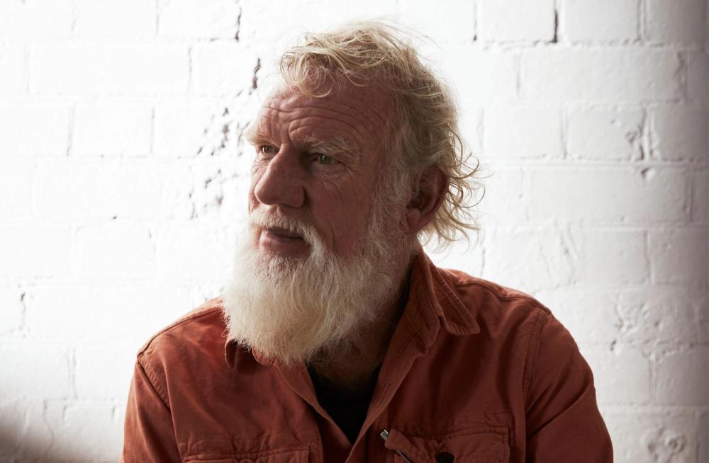 Bruce Pascoe sits in a grey arm chair wearing a peach coloured button up shirt, he has a white beard and is looking upwards and towards the right.