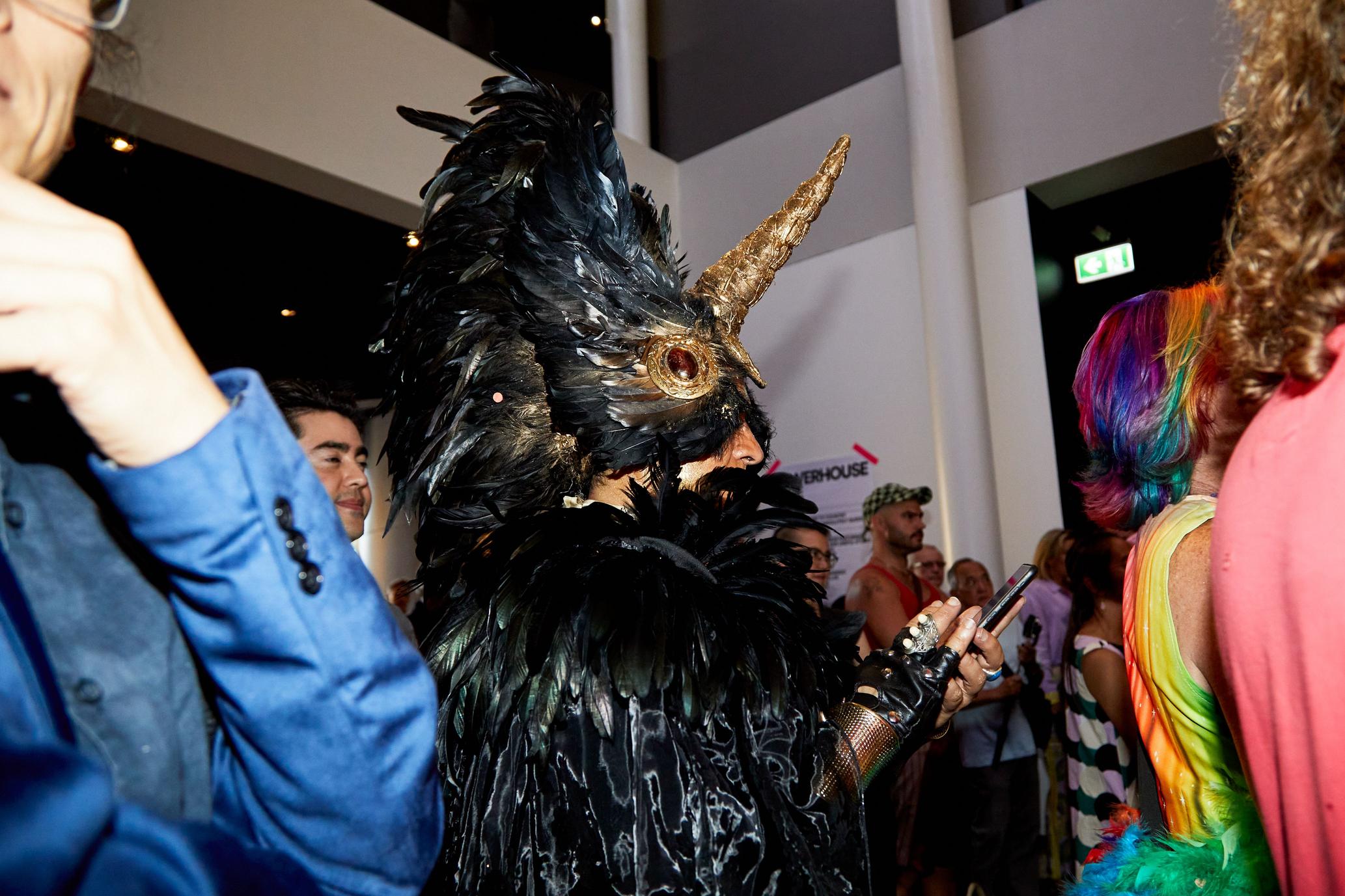 A man stands in a crowd watching a performance. He is wearing an embellished unicorn costume with black feathers, a gold horn and jewels.