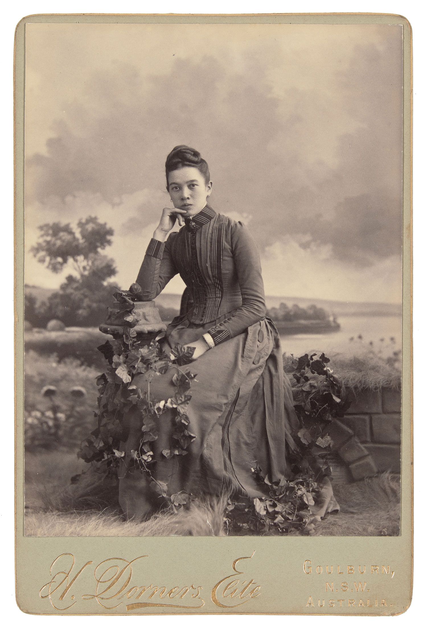 Mounted black and white photograph depicting a girl seated in a long sleeve, full-length dress with foliage on her lap and around her feet.
