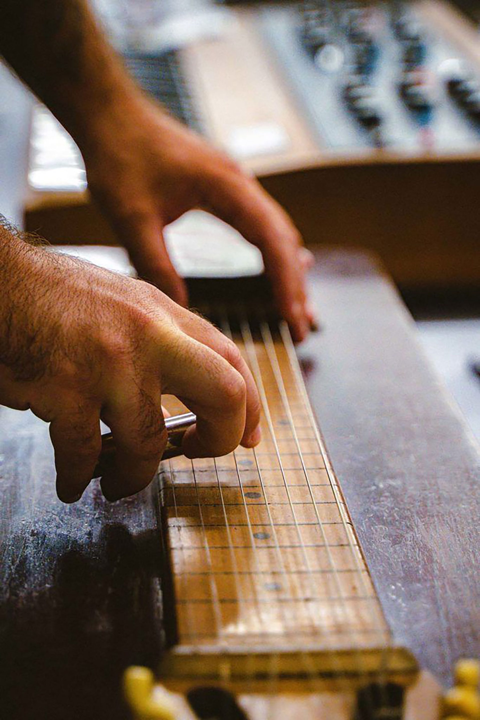 A close up of two hands playing a pedal steel guitar. The left hand is plucking the strings while the right hand is holding a metal slide.