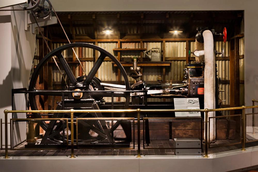 A large horizontal stationary steam engine made of brown metal. A white insulated pipe on the right connects to a boiler and the belt on the left.