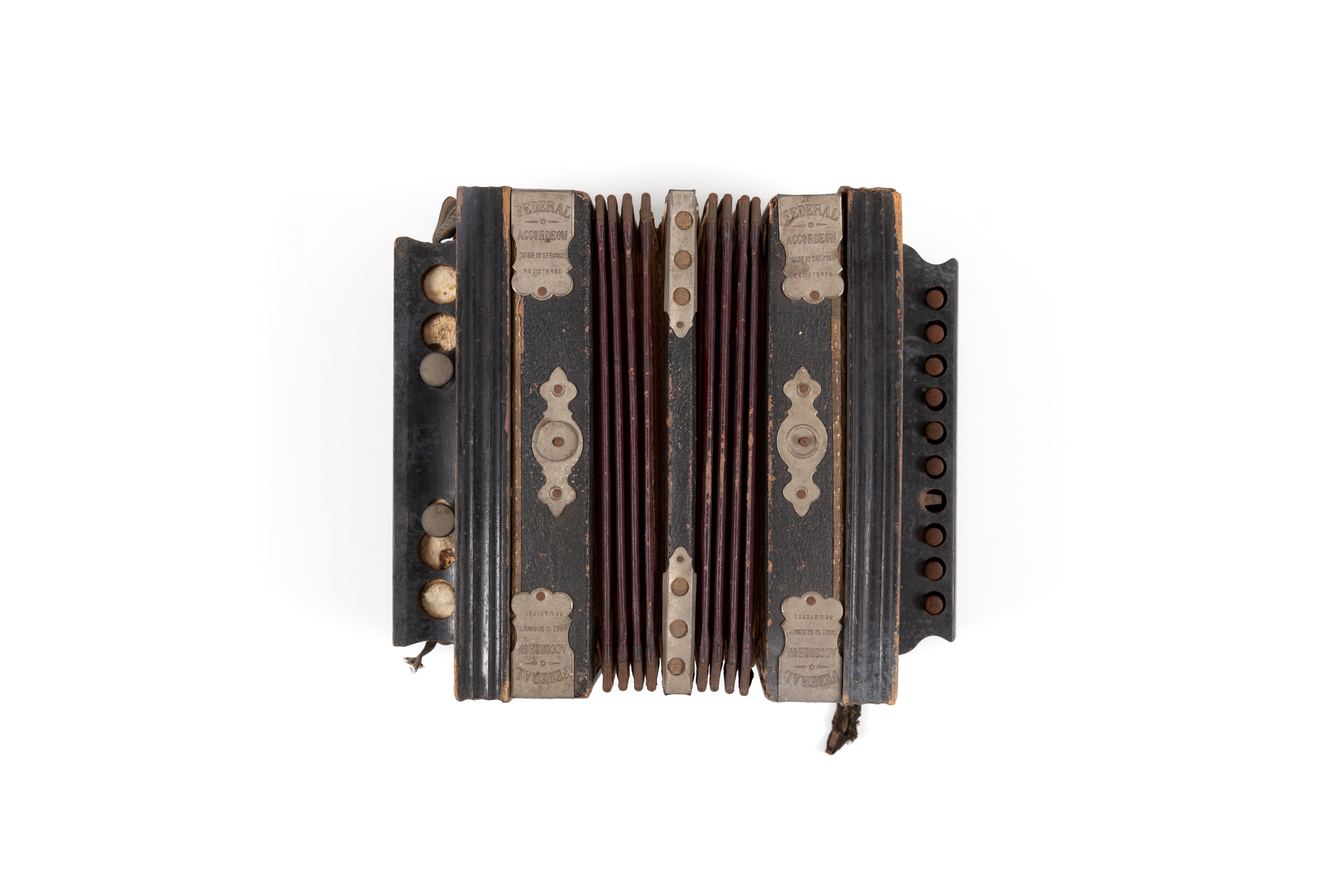 Small musical accordion made from leather, black-painted wood, metal, paper and fabric. Above the keys is the text: "Federal". Metal reinforcing plates on each corner are stamped with the text: "FEDERAL / ACCORDION / MADE IN GERMANY / REGISTERED"