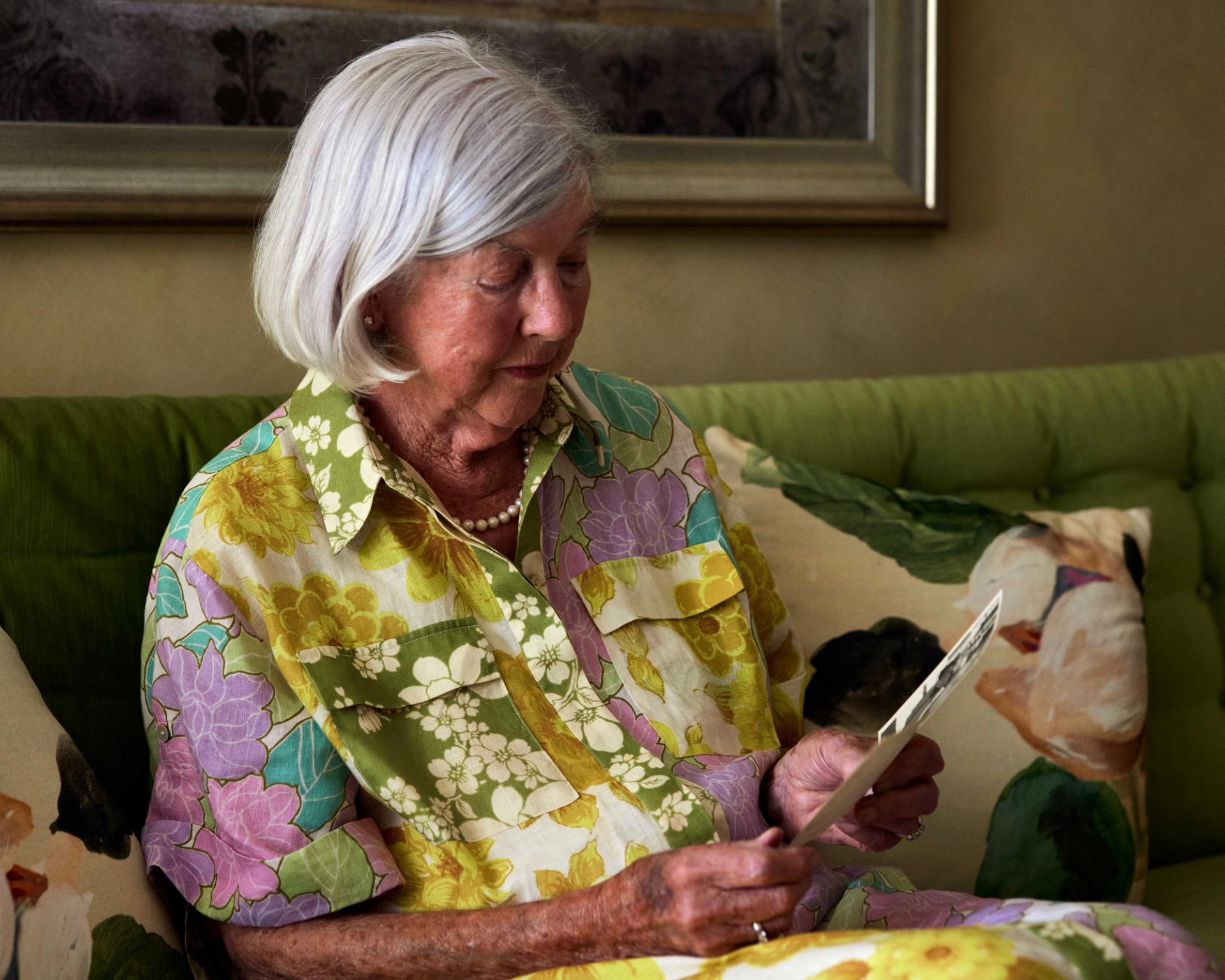 An elderly woman sits on her lounge and looks at two photos she holds in her hands.