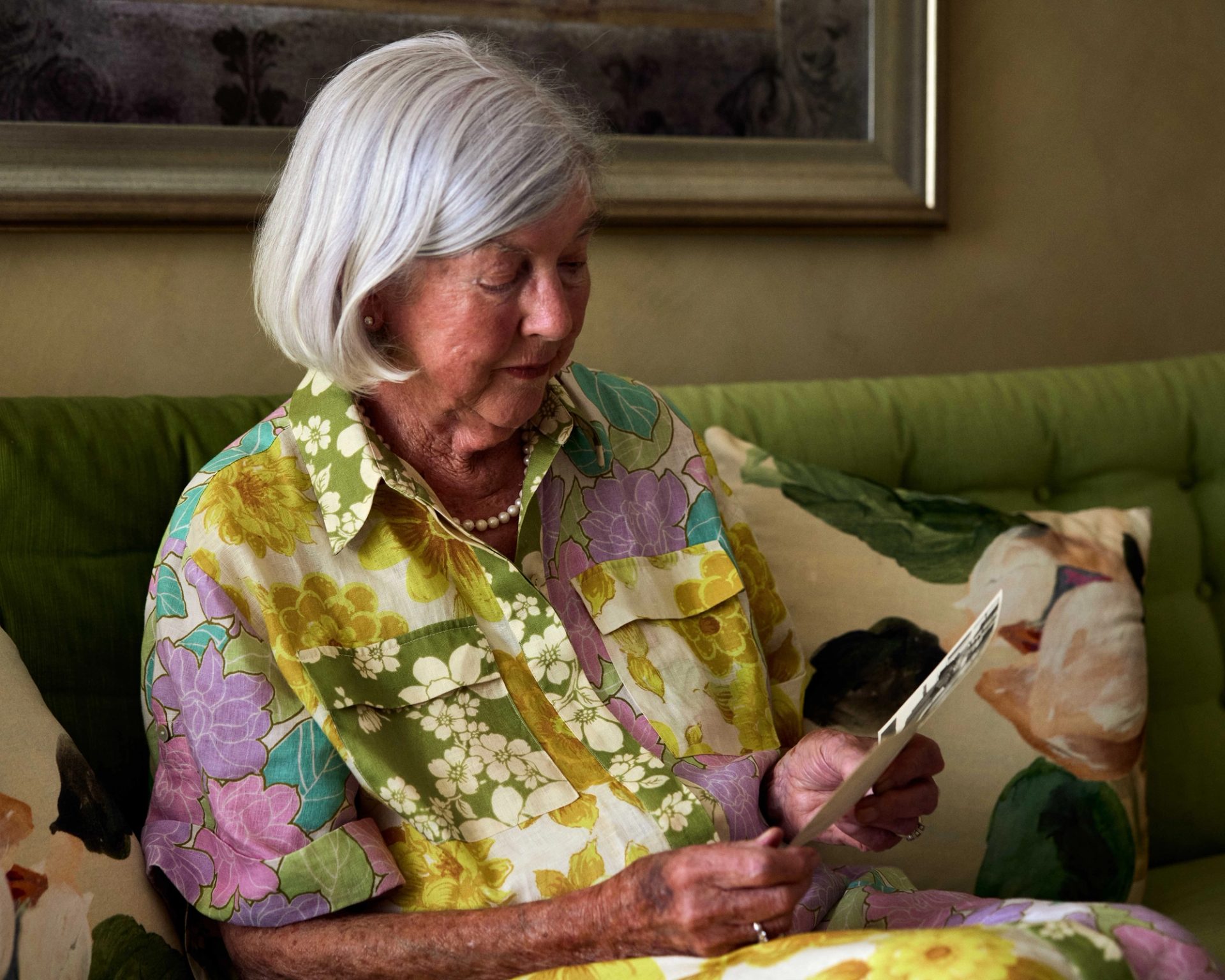 An elderly woman sits on her lounge and looks at two photos she holds in her hands.