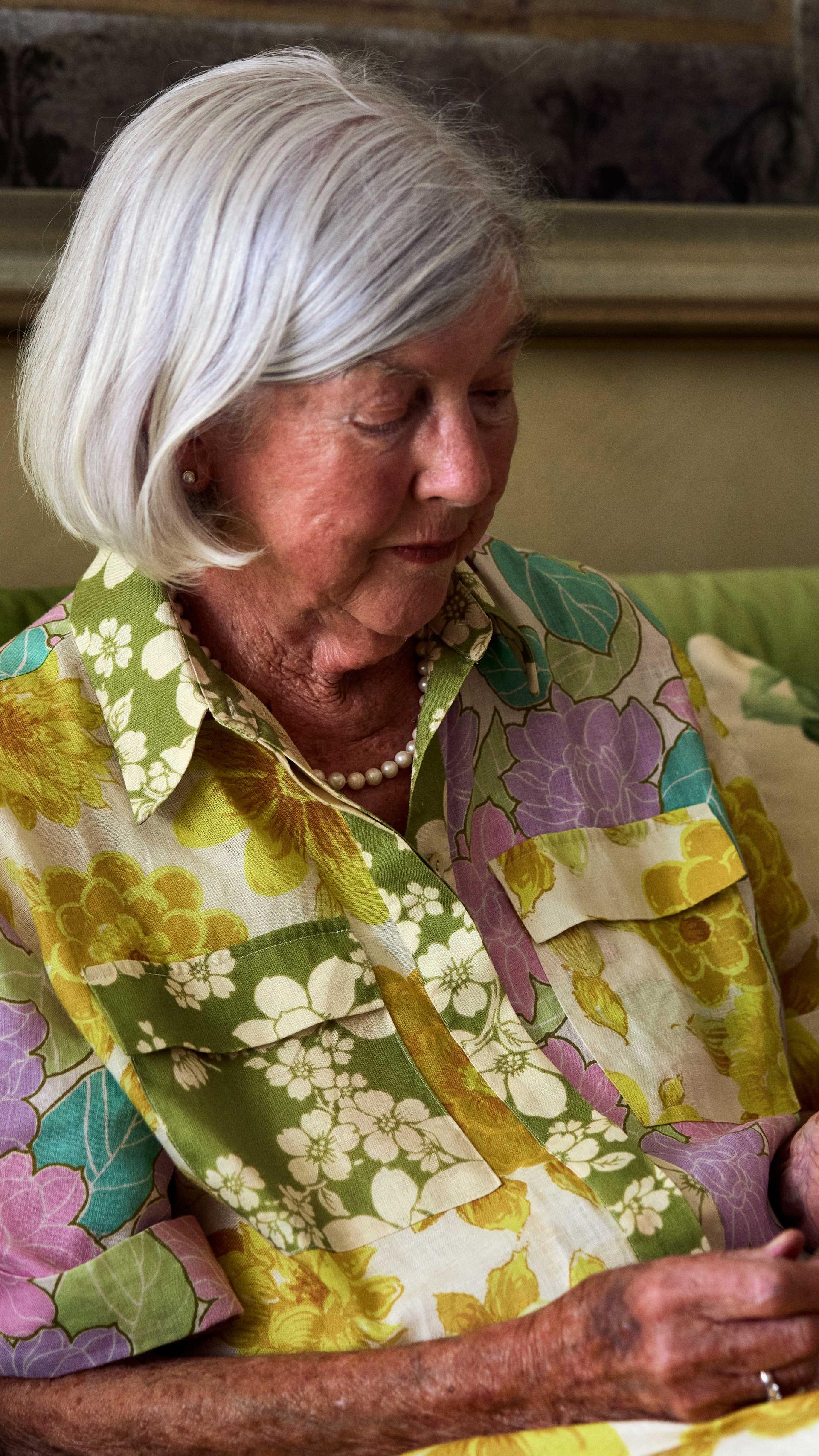 An elderly woman sits on her lounge and looks at two photos she holds in her hands.
