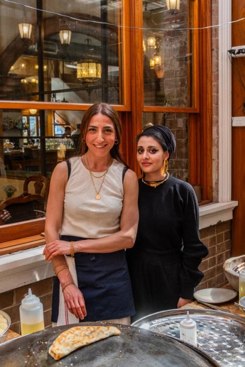 Two women stand together for the camera behind an outdoor kitchen bench.