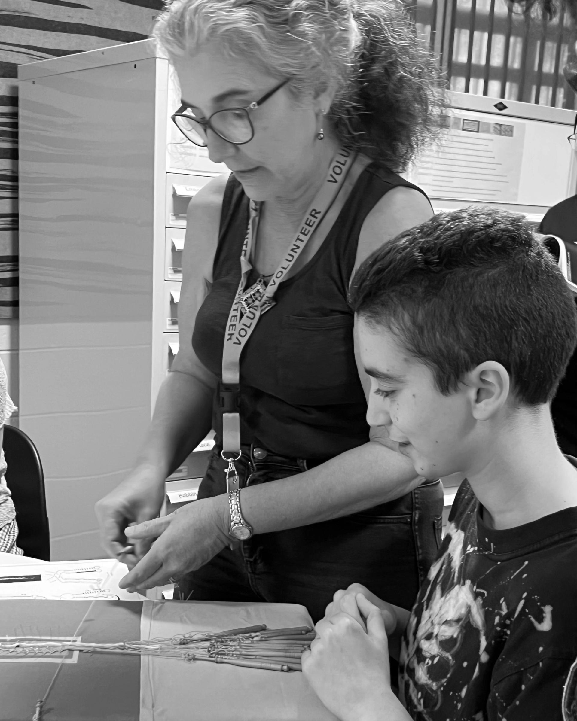 Black and white image of a woman (Isa) teaching a young boy the practice of lacemaking with a lacemaking pillow in front of them.