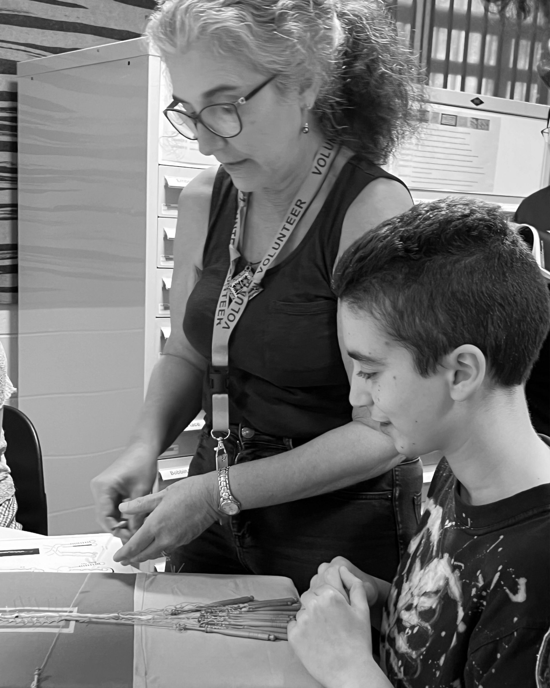 Black and white image of a woman (Isa) teaching a young boy the practice of lacemaking with a lacemaking pillow in front of them.