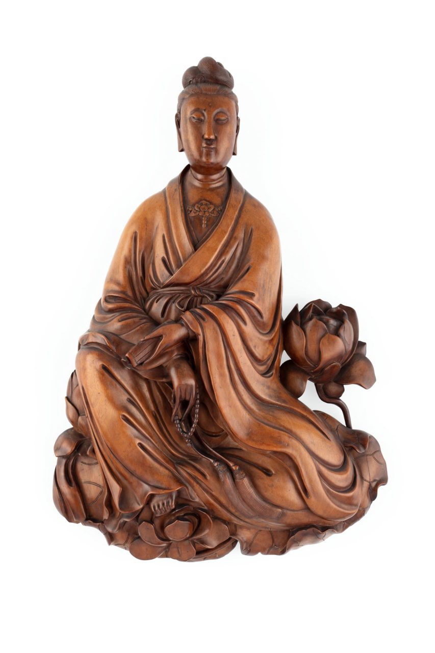 Carved wooden figure of Guan Yin, The Goddess of Mercy, seated with a rosary in one hand and text in the other; she is seated on a lotus leaf and has a lotus flower below her foot and one at each side. The lotus is a symbol of purity and is associated with Guan Yin.