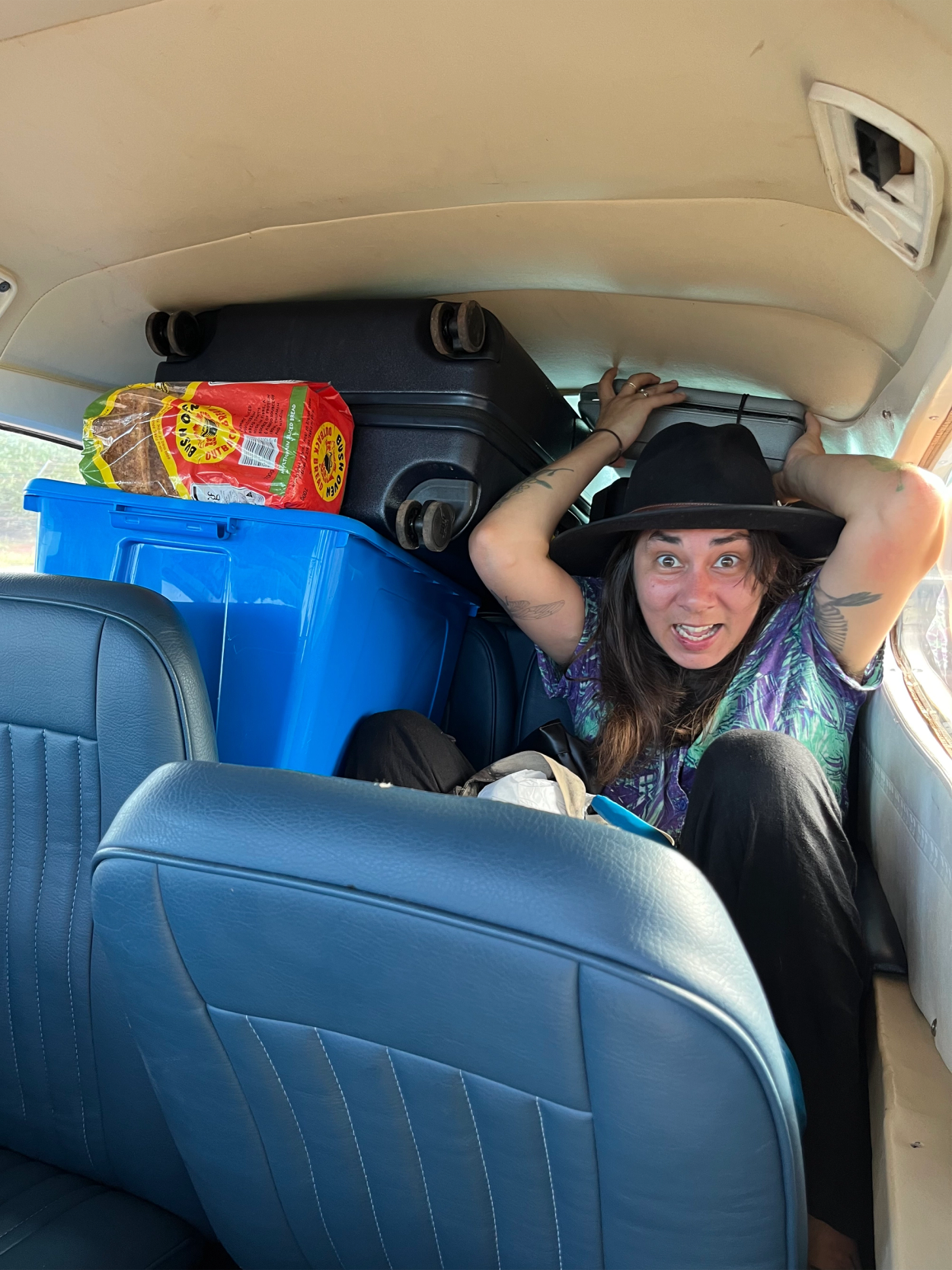 A woman sits in the back of a van with suitcases and bags packed in around her. She has a comical expression on her face.