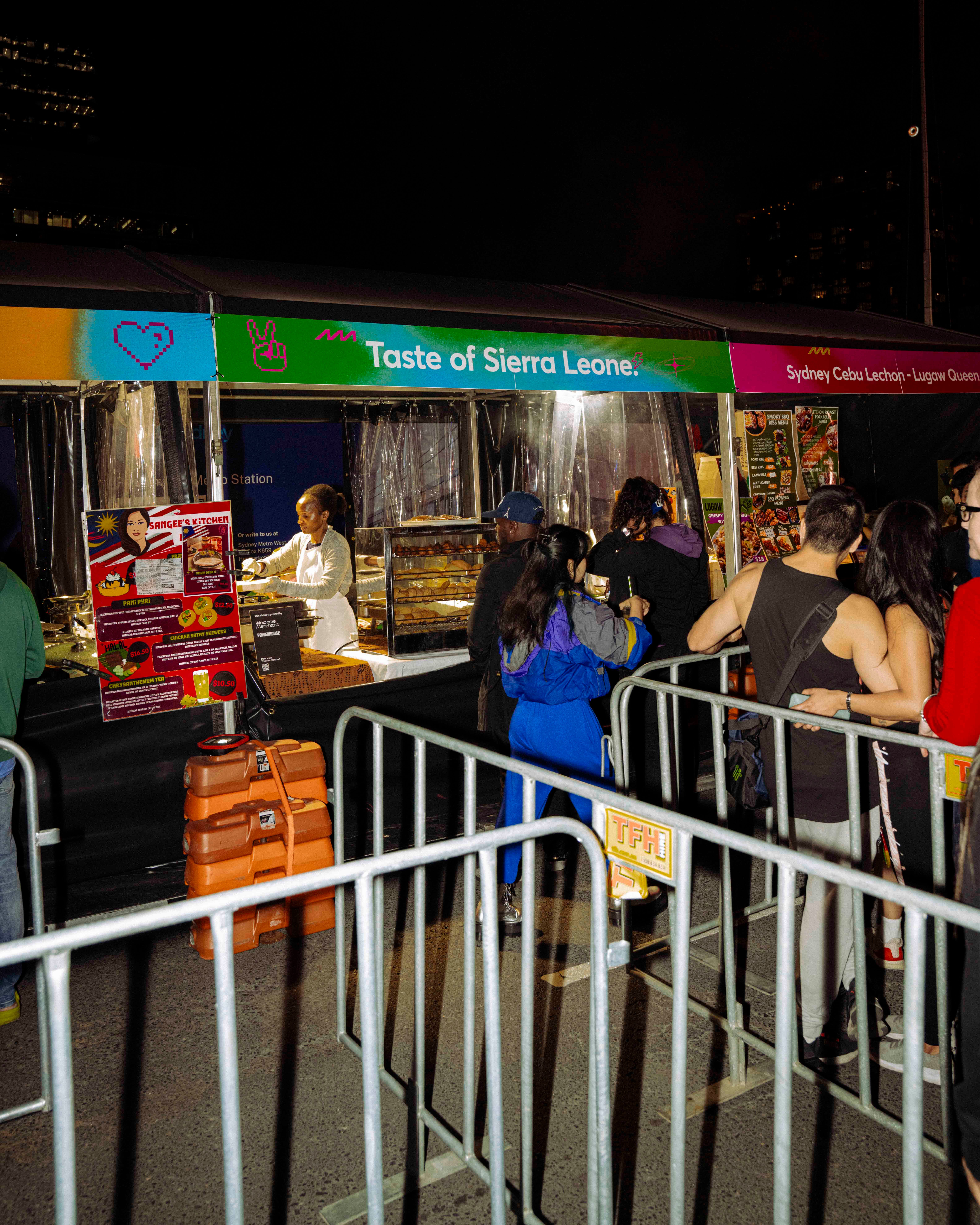 A lively night scene at a busy food stall labelled ‘Taste of Sierra Leone’, with a vendor serving customers who queue along lines of temporary event fencing.