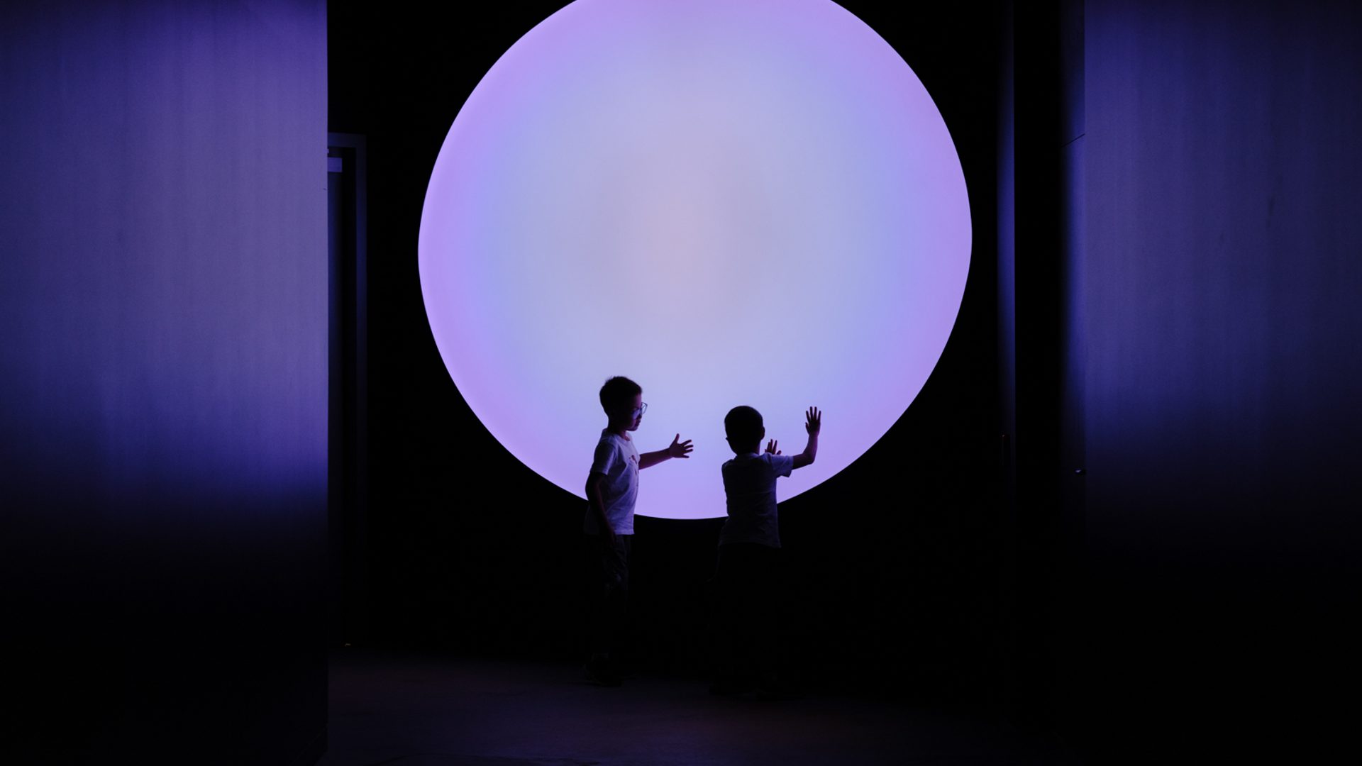 Two young children touch their hands to a big purple circle, that illuminates are dark room.