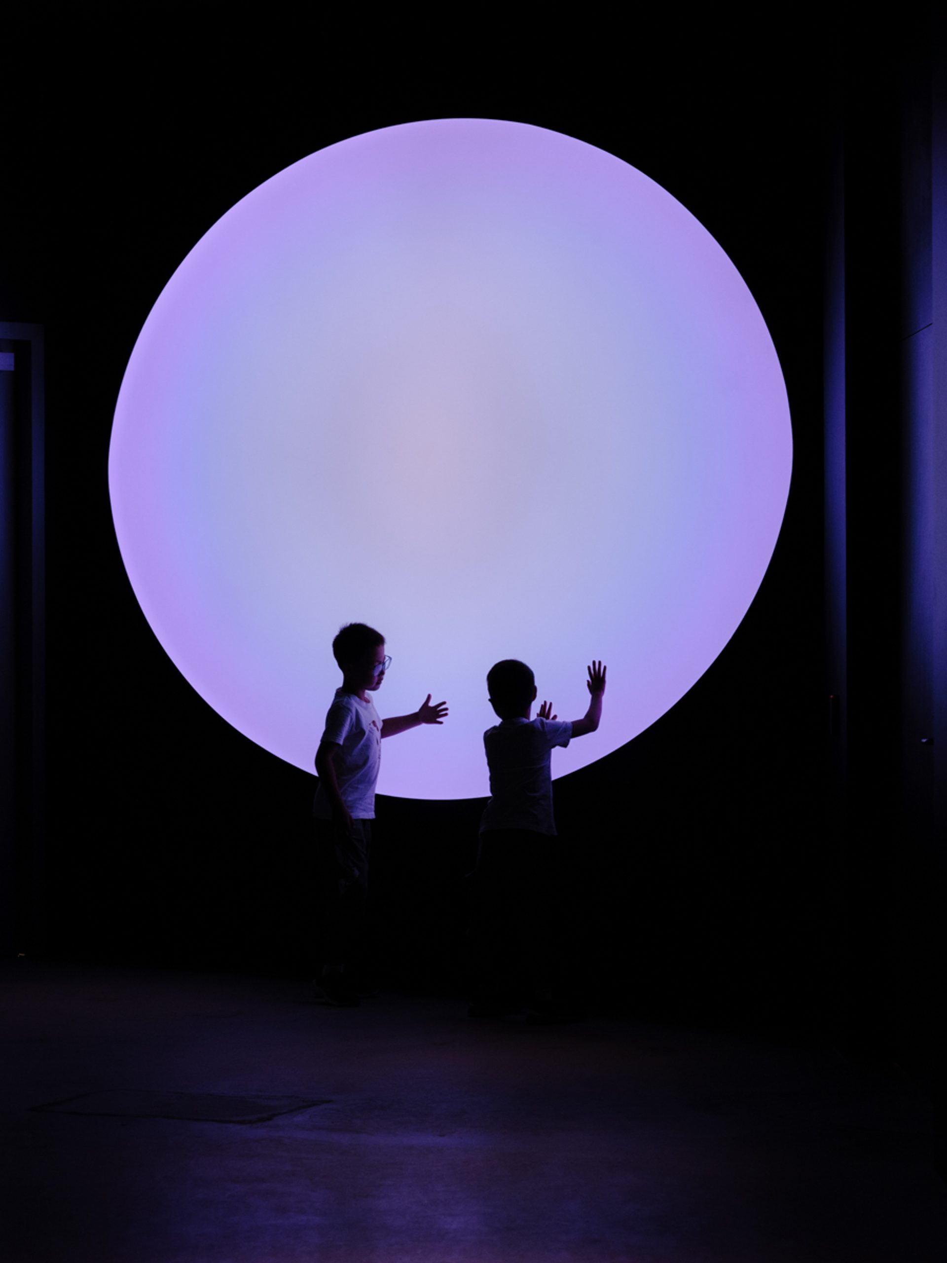 Two young children touch their hands to a big purple circle, that illuminates are dark room.