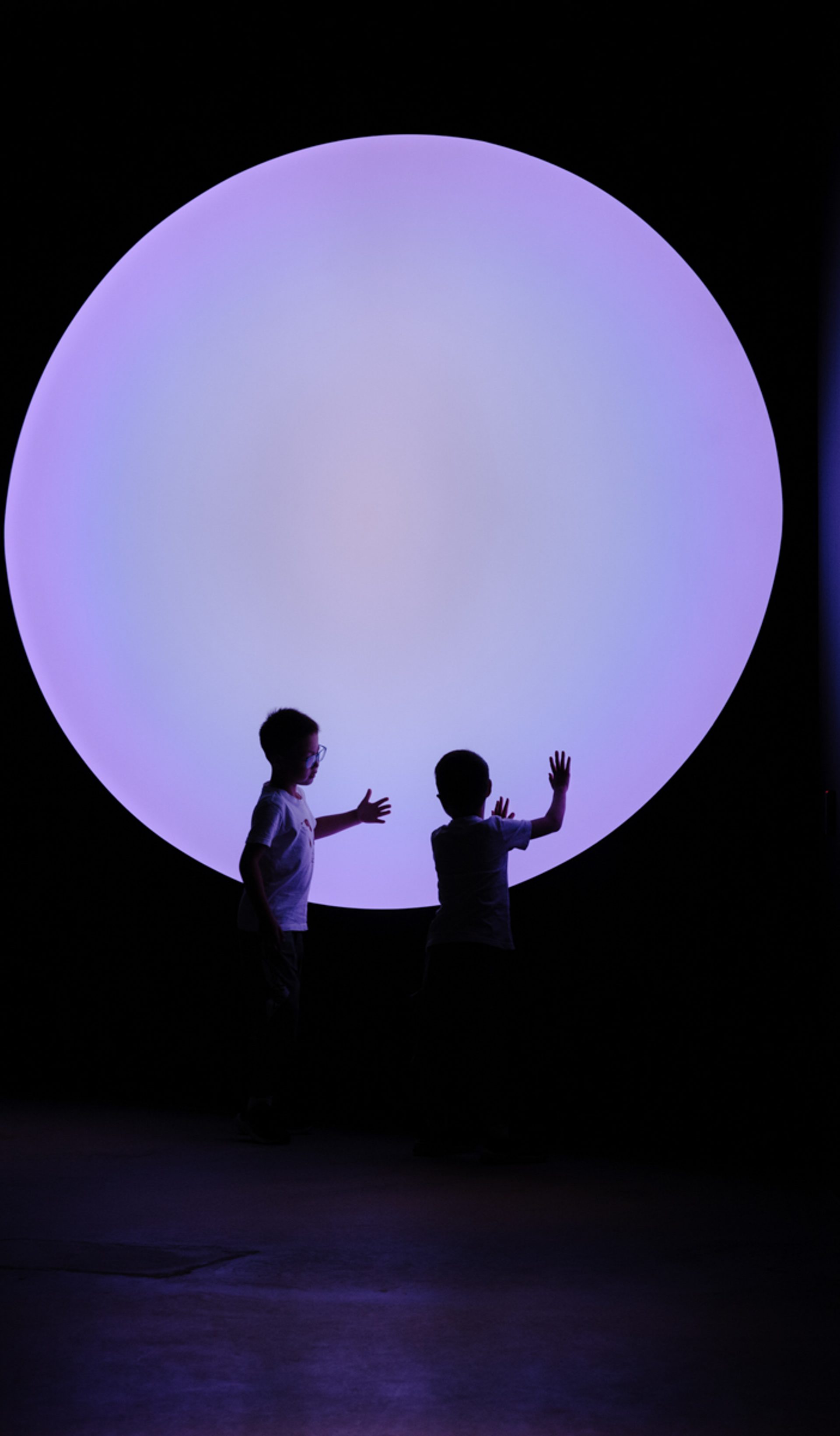 Two young children touch their hands to a big purple circle, that illuminates are dark room.