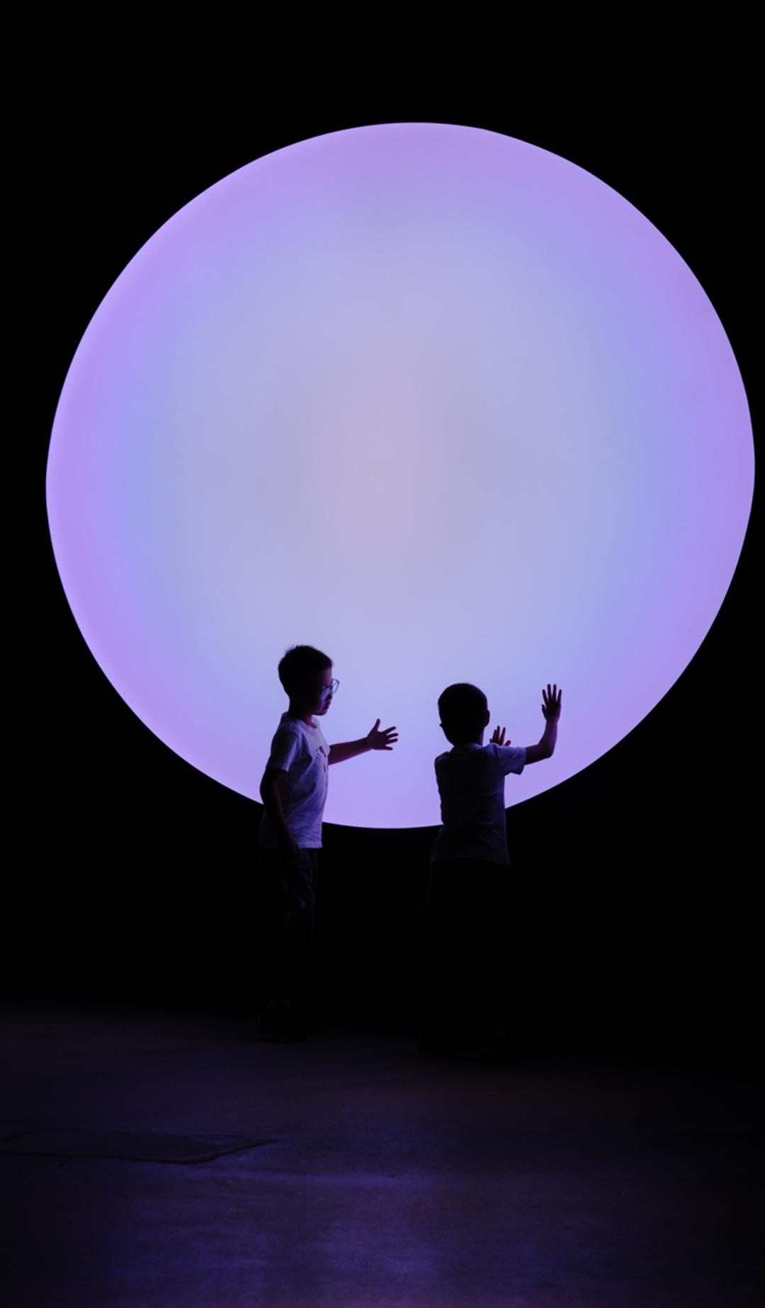 Two young children touch their hands to a big purple circle, that illuminates are dark room.