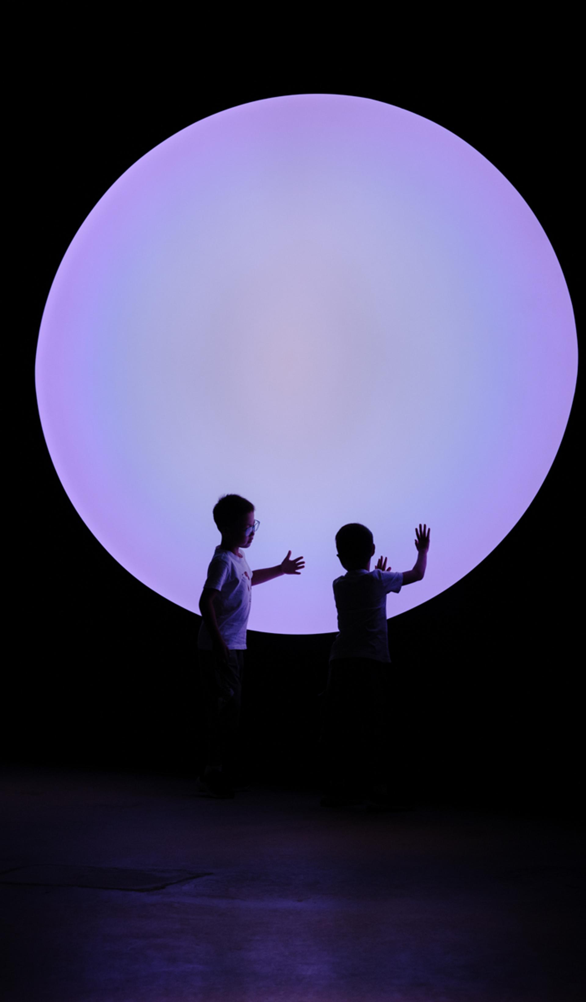 Two young children touch their hands to a big purple circle, that illuminates are dark room.