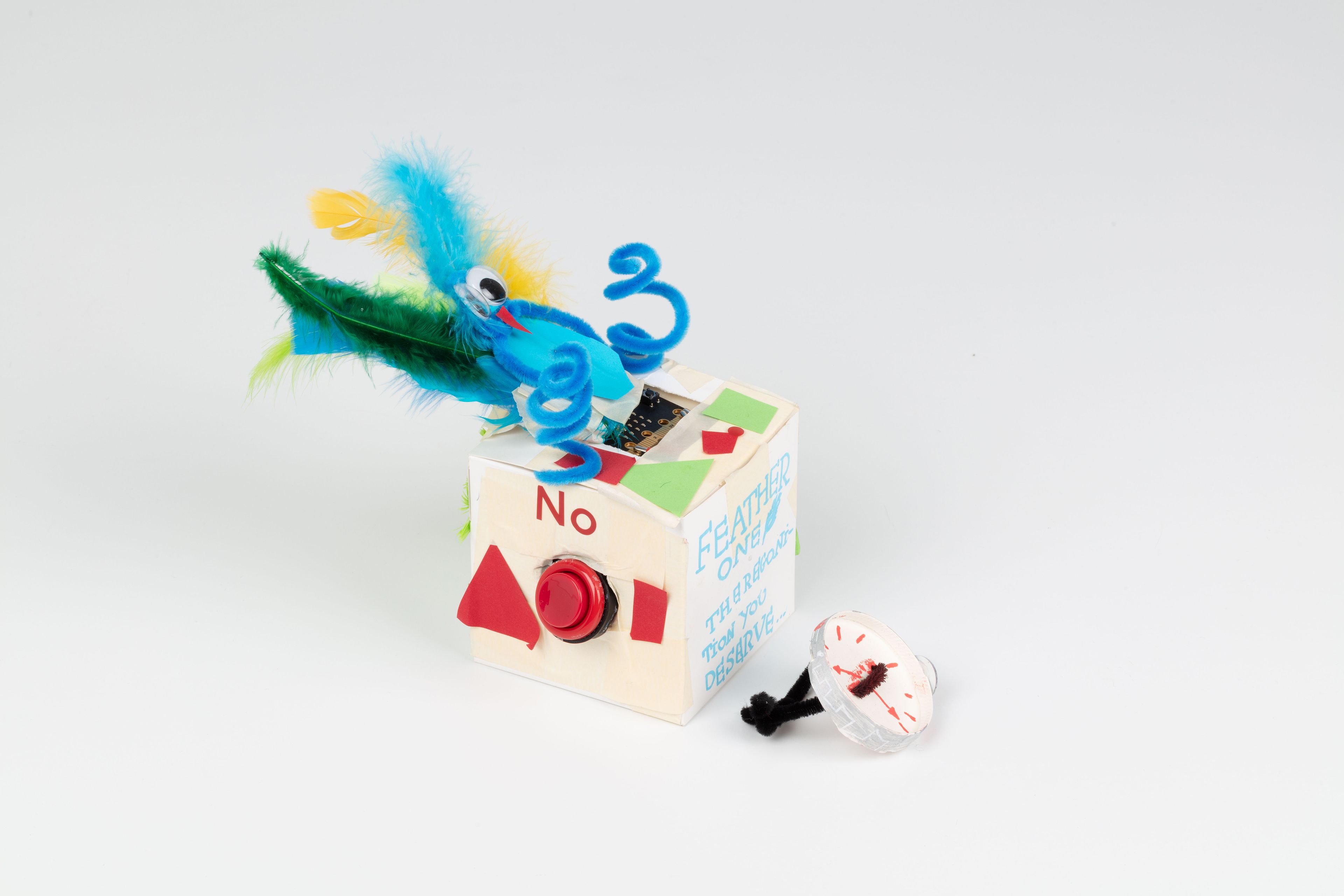Image of a hand crafted mechanical device against a white background. A white cube decorated with cutout paper shapes, feather and pipe cleaners. One side with a red button with the label ‘No’ above it. Beside the cube is a hand crafted watch.