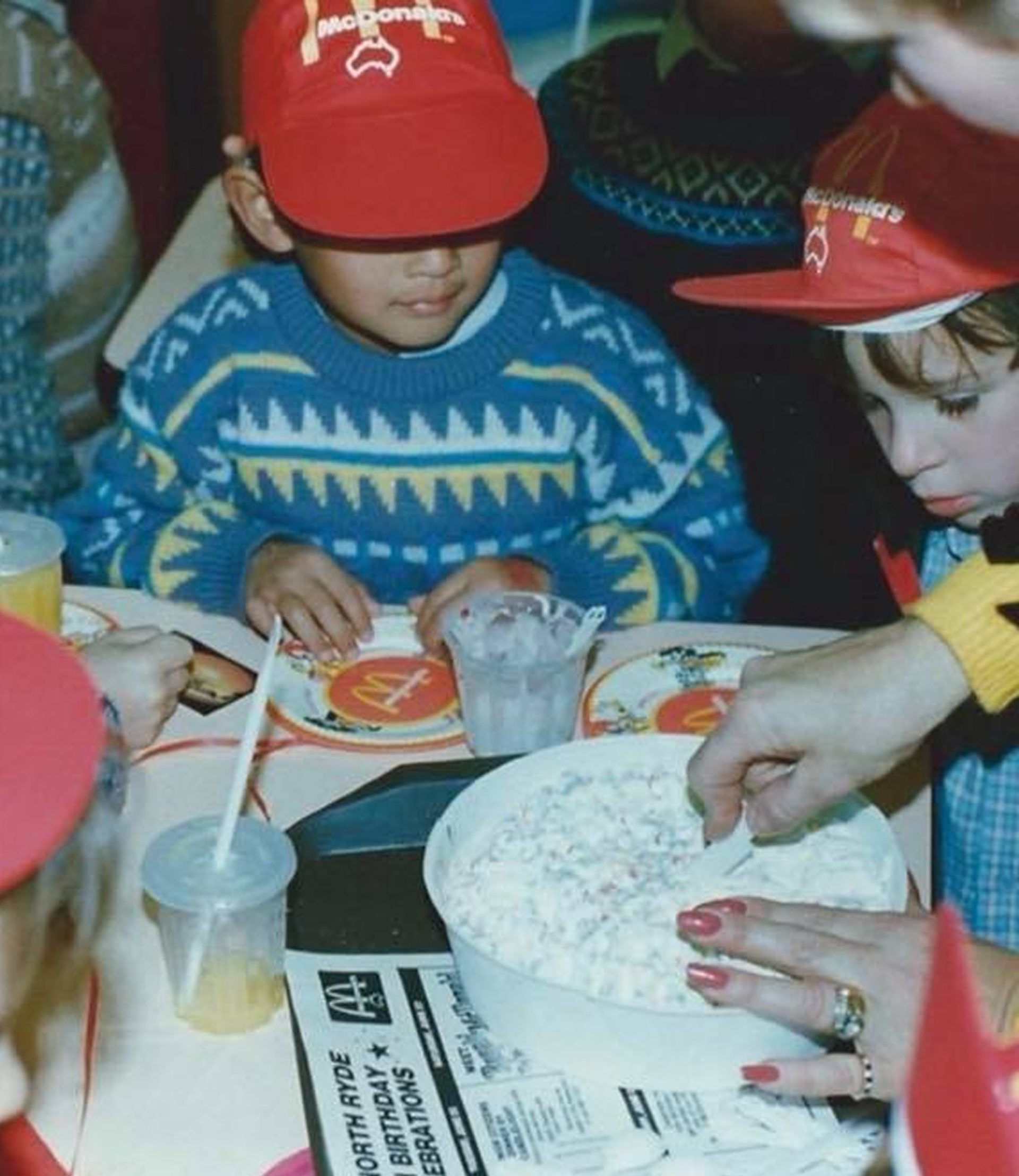 A photograph of kids at a McDonald's birthday party, wearing red hats and looking down to an adult cutting a cake.