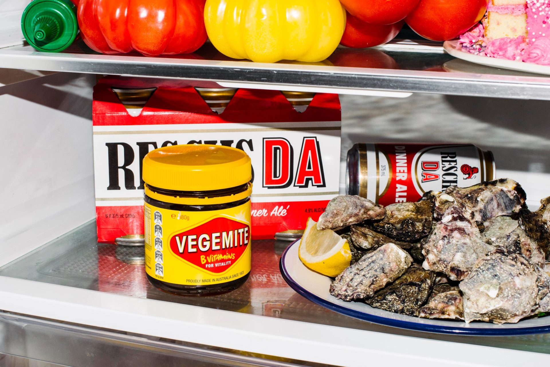 A jar of Vegemite sits on a fridge shelf. Behind it is a six pack of Resch’s beer.
