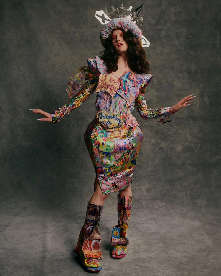 A model poses against a grey background wearing a multicoloured structured dress and metal crown